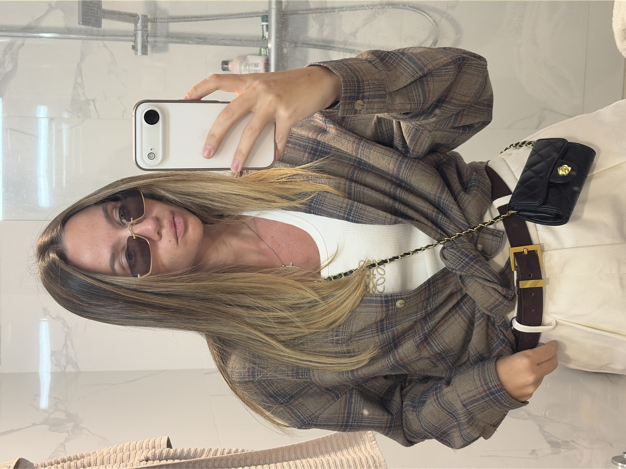 A woman is taking a selfie in a bathroom. The background includes a showerhead and a white marble wall. She is wearing sunglasses, a white shirt, a brown plaid shirt tied at the waist, cream-colored pants with a brown belt, and a small black quilted purse with a gold chain. She is holding a white cell phone. The location is in the city of Unknown, Unknown.