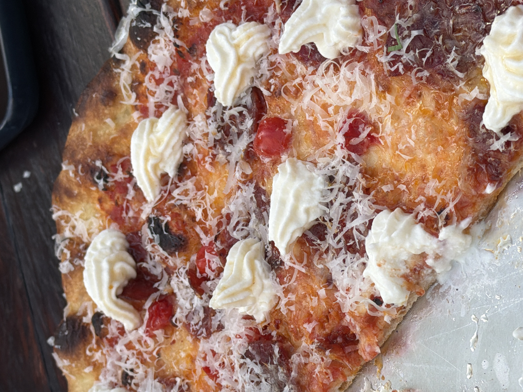 The image displays a close-up view of a partially eaten pizza on a metallic serving tray. The pizza has a browned crust, suggesting it was baked in a high-temperature oven, with darkened areas indicating slight charring. The toppings appear to include tomato sauce, shredded parmesan cheese, dollops of a creamy white topping (possibly ricotta or a similar cheese), and what seems to be roasted red peppers or cherry tomatoes. Some of the crust has been eaten, revealing the metallic tray underneath. The image is shot from a high angle, looking down onto the pizza. Part of a dark object, potentially a mug or cup, is visible in the upper left corner of the frame. The composition emphasizes the textures and colors of the pizza toppings and crust. There is no information available in the image to suggest a location in the city of London, United Kingdom.