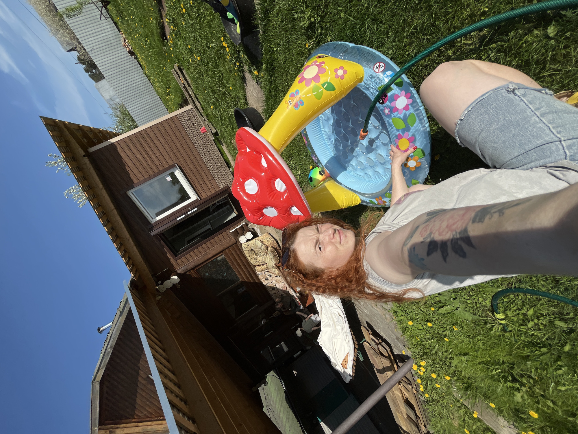 A medium-shot outdoor selfie shows a fair-skinned woman with long, wavy, reddish-orange hair and a tattoo on her left arm. She is reclining on a green lawn dotted with yellow dandelions, with her right knee visible in blue denim shorts. In the background, a small, wooden cabin-like structure with a brown-tiled roof and a window is visible, along with the blue sky. Next to her is an inflated plastic kiddie pool, partly filled with water, and a green garden hose filling it. Next to the pool is an inflated red mushroom decoration and a yellow arch. Based on the location, Chaikovsky, Russia, and the architecture, this is likely a private residence or a vacation home outside the main city.