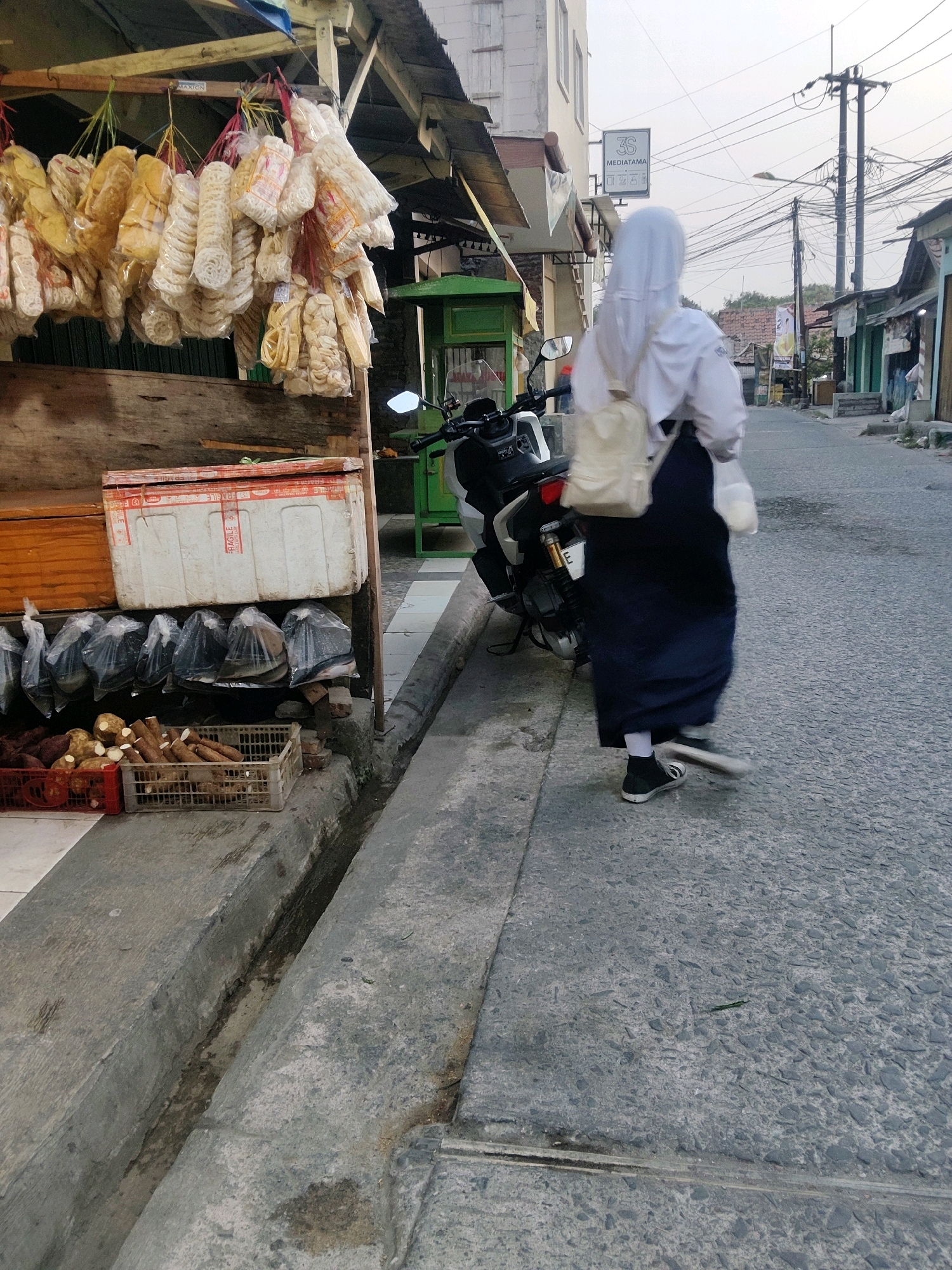 In the city of Mekarmukti, Indonesia, an individual wearing a white headscarf, white long-sleeved shirt, and a dark blue skirt walks away from the camera on a paved road. The person carries a light-colored backpack and a white plastic bag. They are walking past a sidewalk lined with various stalls. In the foreground, a stall displays an assortment of bagged goods and produce, including what appears to be tubers and root vegetables in baskets. A white styrofoam cooler is also visible on the stall. Behind the person, a motorcycle is parked. The background shows buildings, power lines, and more street stalls, suggesting a marketplace or a busy street scene. The lighting indicates it is either early morning or late afternoon.