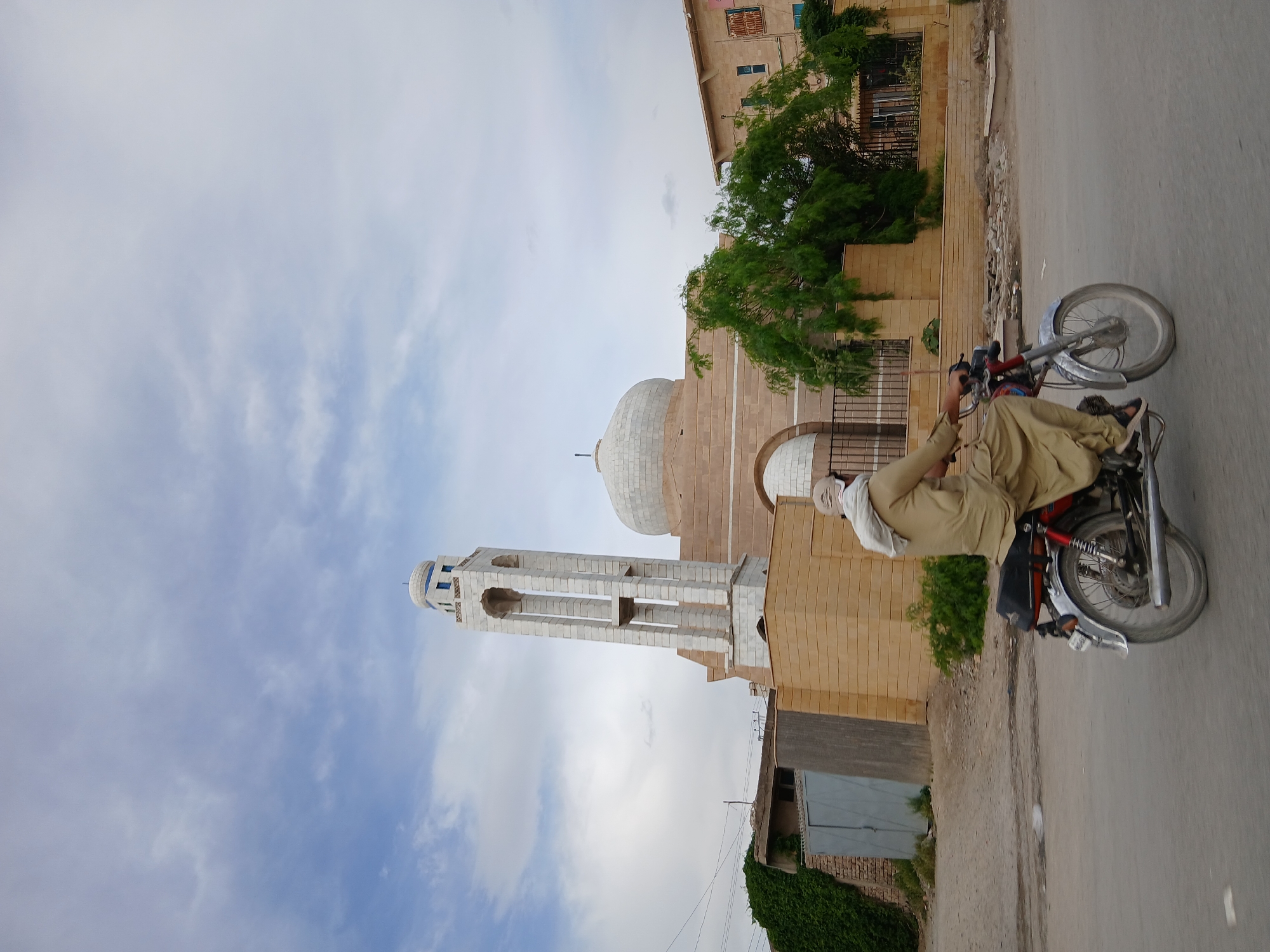 A street-level photograph shows a motorcyclist with a mosque in the background. The man is riding a red motorcycle on a paved road. He is wearing light brown clothing and a white head covering. The mosque has a tall minaret and a dome, constructed of light beige bricks. The sky is light blue with scattered white clouds. The location appears to be Quetta City Tehsil, Pakistan, based on architectural styles typical of the region. Other buildings are present in the background along the street.