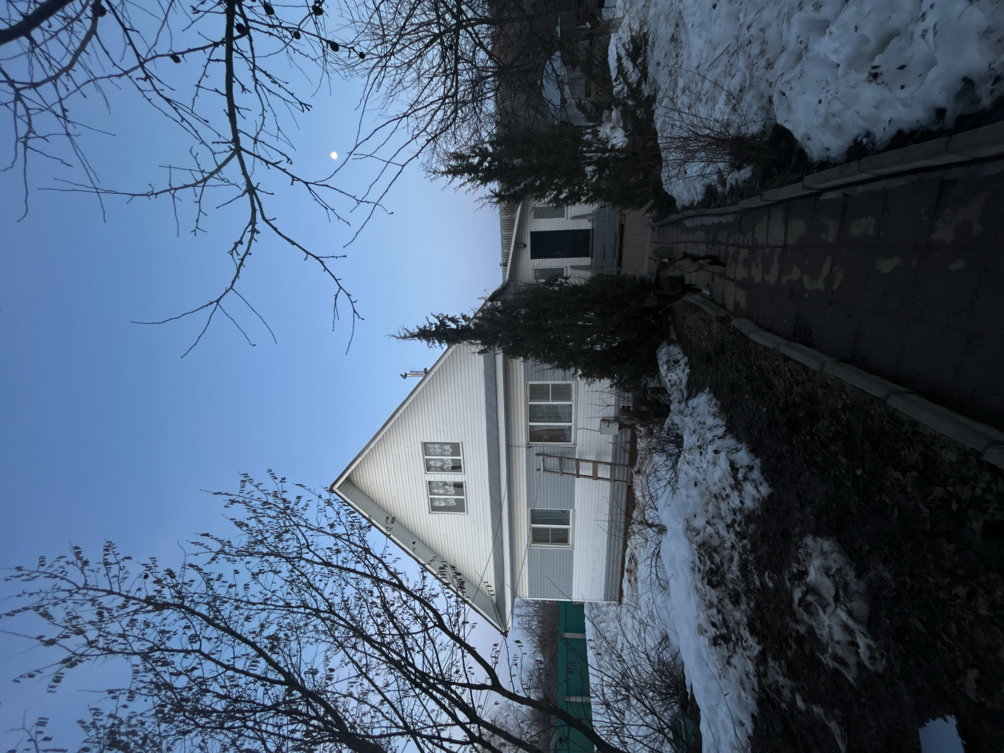 The image shows a two-story house in Фоки, Russia. The house has a white upper story and a gray lower story. A ladder leans against the side of the house. There are trees in the foreground and background, and snow is visible on the ground. A path leads to the front door. The sky is blue, and a small moon is visible.