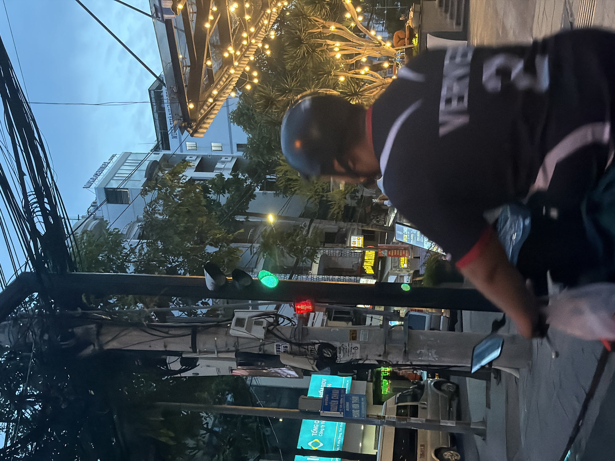 The image depicts a street scene in Nha Trang Ward, Vietnam. A person wearing a helmet and a black shirt with the word "VERVEL" on the back is on a scooter. A traffic light with green and red lights is visible in the background. Utility poles with numerous wires are on the left side of the image. There are buildings with signs in Vietnamese. There are trees decorated with string lights.