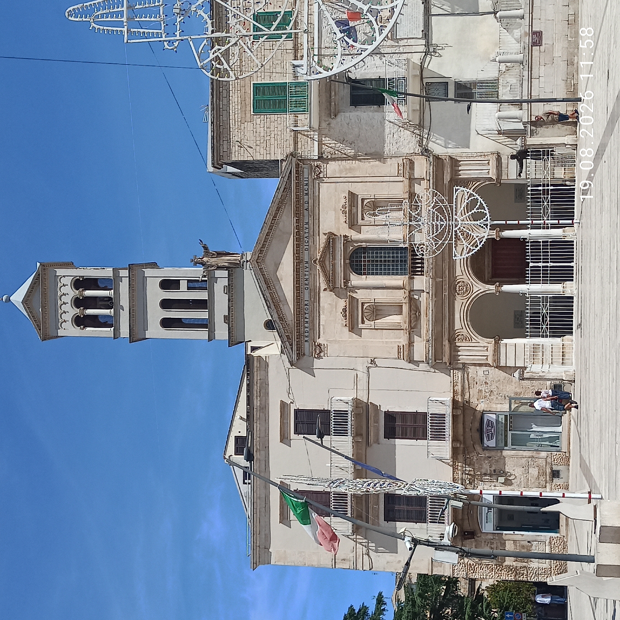 The image shows a full frontal view of the Basilica Cattedrale di Santa Maria Assunta in Ruvo di Puglia, Italy. The cathedral features a prominent bell tower with arched openings and a statue of Jesus Christ at the top. The facade of the cathedral is adorned with inscriptions and architectural details. An Italian flag flies from a pole to the left of the cathedral.
In the foreground, a paved public square is visible with people walking. Some are dressed casually, suggesting a warm day. The square is equipped with streetlights and some decorative light installations, possibly for a festival or event. A date stamp "19.08.2026 11:58" is overlaid on the lower right portion of the image, indicating the time and date of capture. The sky is clear and blue. The surrounding buildings are made of stone and stucco, with traditional shutters on the windows.