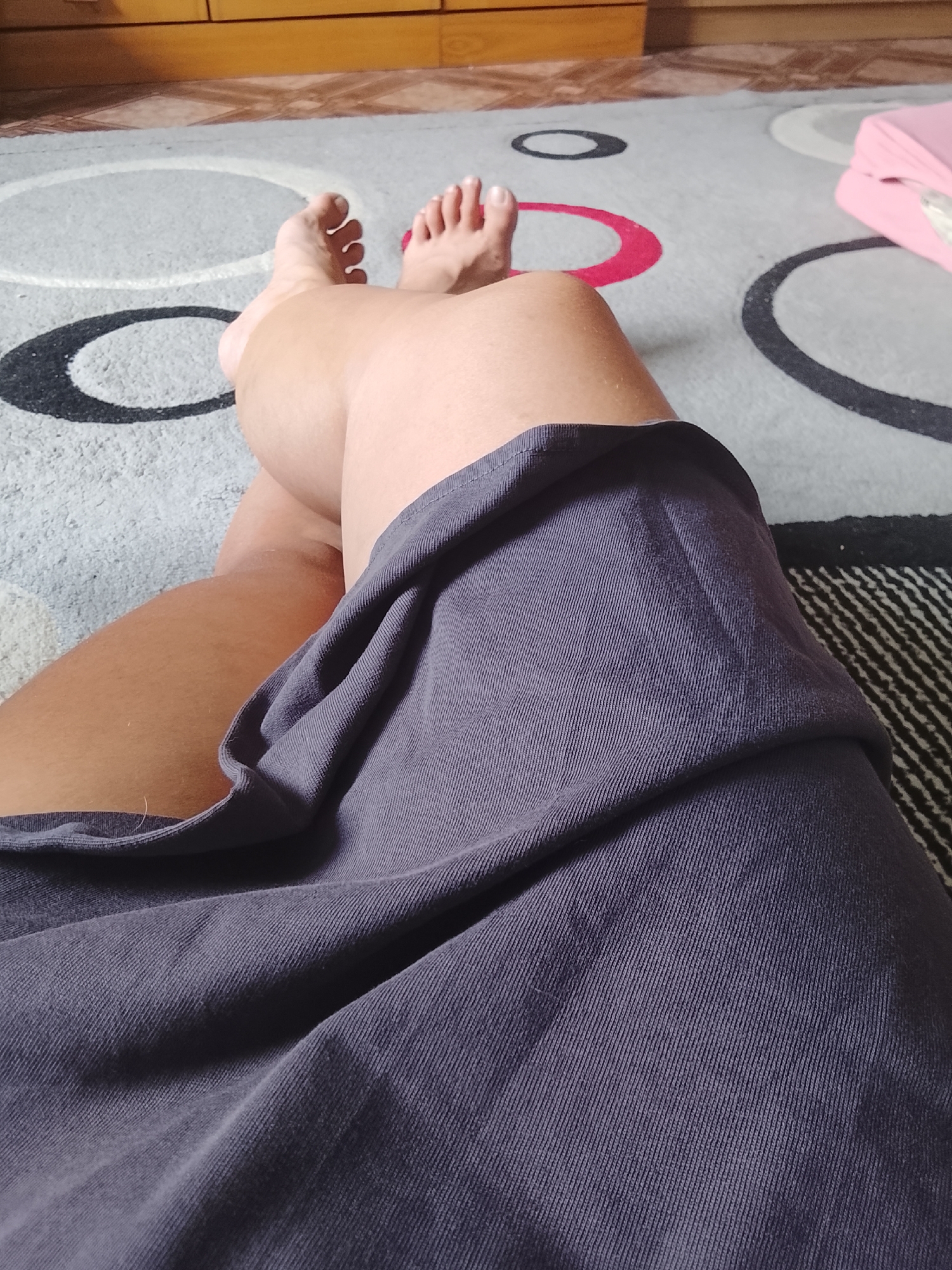 The image shows a close-up view of a person's legs, resting on a carpet with a geometric pattern of circles in shades of gray, black, and red. The person is wearing a dark gray skirt. The background consists of a wooden floor and a piece of furniture, possibly a dresser, in a muted brown color. A pink object, which appears to be a cushion or a small bag, is partially visible on the right side of the image, resting on the carpet. The lighting suggests an indoor environment. There are no discernible elements in the image that would allow for identification of the specific location within Almaty, Kazakhstan.