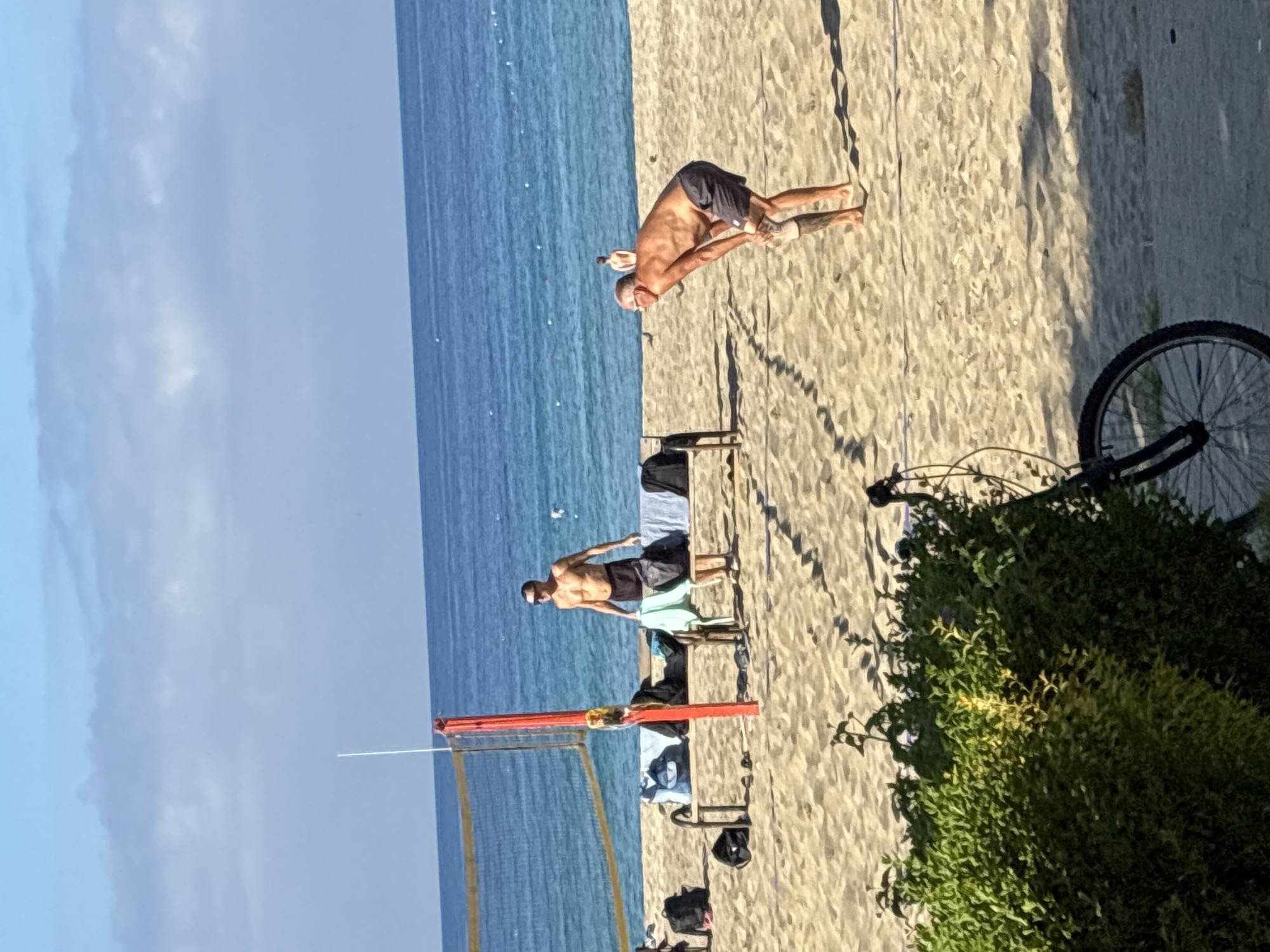 The image, when viewed upright, captures a sunny day at a beach in Alanya, Turkey, showing the sea, sand, and sky. The vast blue ocean occupies the left side of the frame, meeting a sandy beach on the right. Above, the sky is clear and blue with scattered white clouds.
Two men are visible on the beach. In the right mid-ground, a shirtless man wearing dark shorts, who appears bald and is wearing sunglasses, is bending forward, possibly stretching or engaging in a physical activity. Further left, near the center of the frame, another shirtless man in dark shorts stands near a red-posted beach volleyball net, facing the ocean. He also appears to be wearing sunglasses.
Several beach chairs or loungers are scattered around, some with clothes or towels draped over them. A partially visible bicycle, specifically its front wheel and handlebars, is seen on the far right, situated among lush green bushes and vegetation that line the edge of the beach.
The bright, direct sunlight casts long, well-defined shadows across the sand, indicating a clear, sunny day, likely morning or late afternoon. The overall scene suggests a leisurely and recreational atmosphere on the beach. No visible text is present in the image.