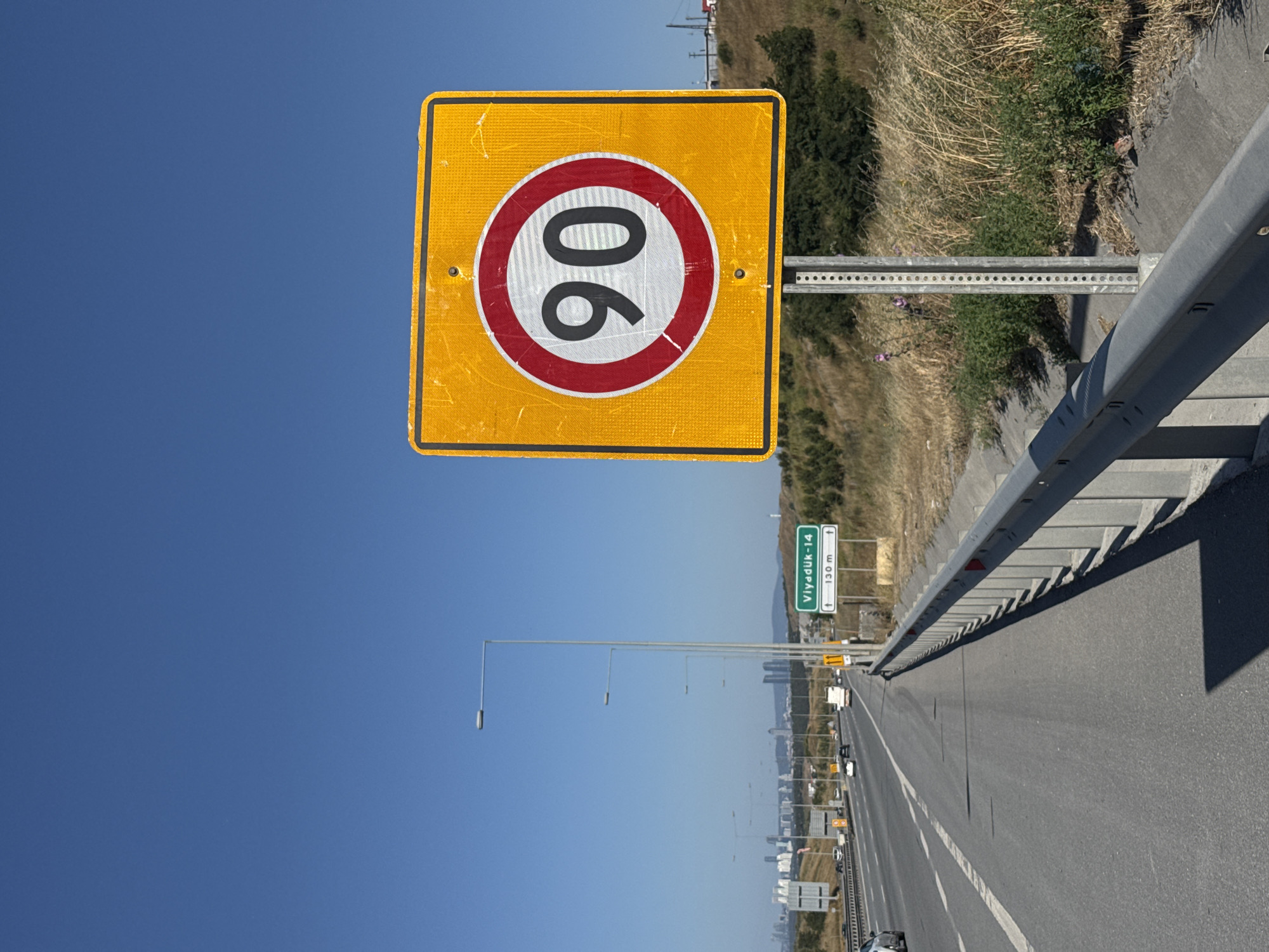 A bright yellow, square speed limit sign with a black border is prominently displayed against a clear blue sky. The sign features a circular red border with a white inner circle, containing the number "90" in black digits. The sign is mounted on a metal pole.
The scene depicts a highway, with a grey metal guardrail running along the right side of the road. The highway is asphalt and appears to be in good condition, with white lane markings visible. On the left side of the highway, there is a slope covered with dry grass and some green bushes, suggesting a rural or semi-rural setting. In the distance, buildings and possibly city structures are visible, indicating a populated area. A green road sign with white text "Viyadük-14" and "130 m" with arrows pointing forward is also visible, along with several lampposts lining the highway. Several vehicles, including cars and trucks, are on the highway, suggesting ongoing traffic. The image appears to have been taken during the daytime, with bright sunlight casting shadows. The overall impression is of a clear, sunny day on a Turkish highway.