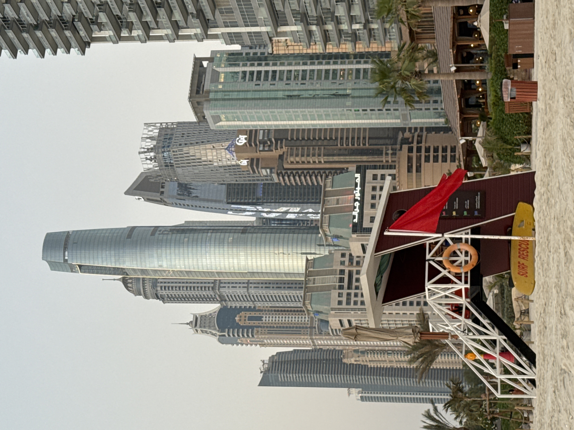 This image depicts a beach scene in Dubai, United Arab Emirates, with a backdrop of modern skyscrapers. In the foreground, a red and white lifeguard tower stands on a sandy beach. A life preserver ring hangs on the tower's railing, and a yellow surfboard with "SURF RESCUE" written on it lies on the sand nearby. A red flag is displayed on the tower. Palm trees and beach umbrellas are scattered around the tower, suggesting a resort or public beach area. Behind the beach, a dense cluster of various skyscrapers, characteristic of Dubai's iconic skyline, rises into the hazy sky. The architecture of the buildings ranges from sleek, glass-covered towers to more ornate, older structures. Some buildings appear to be residential or hotel complexes, while others are clearly commercial or office buildings. The overall atmosphere suggests a sunny, warm day, though the sky is overcast, possibly indicating a light haze.