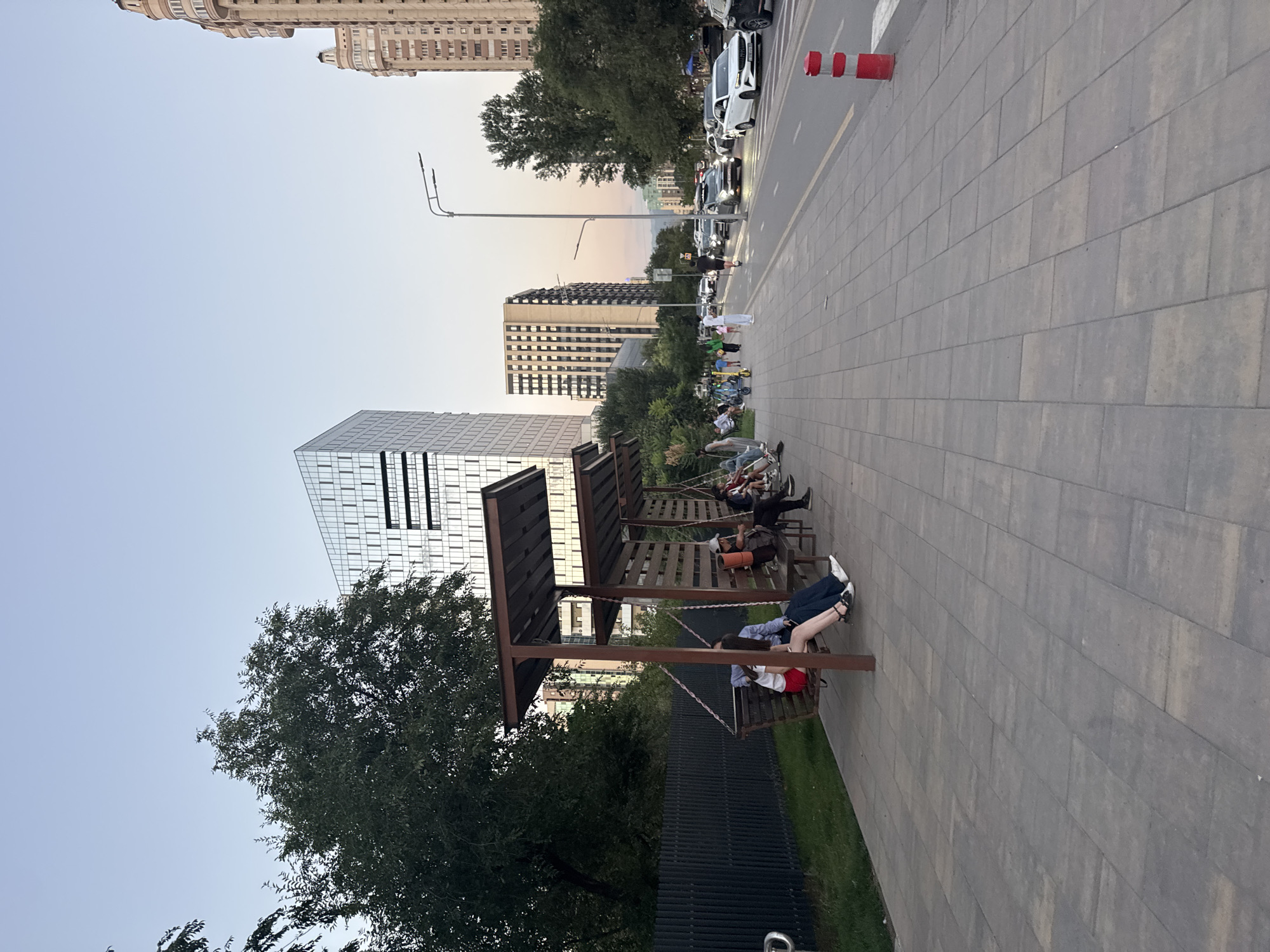 The image depicts a street scene in Almaty, Kazakhstan, likely during the evening given the soft lighting. A pedestrian walkway, paved with rectangular grey tiles, runs along a street with moderate traffic. On the left side of the walkway, there is a series of covered benches and swings, some of which are occupied by people. One couple is seated on a swing bench, with the woman wearing a white top and red shorts, and the man in a blue shirt and dark pants. Further down the walkway, other individuals are seated on benches, some with bags or belongings.
Adjacent to the walkway is a grassy area and a dark metal fence. Beyond the fence, trees and vegetation are visible. The background features modern city architecture, including a distinctive glass-fronted skyscraper with a geometric design and other multi-story buildings. A street lamp is visible above the walkway. On the street, cars are driving, including a white sedan and several other vehicles. A red and white traffic cone is positioned near the curb on the right side of the image. Several people are walking along the pedestrian path, including individuals on scooters. The sky above is clear and a pale blue, suggesting a clear evening.