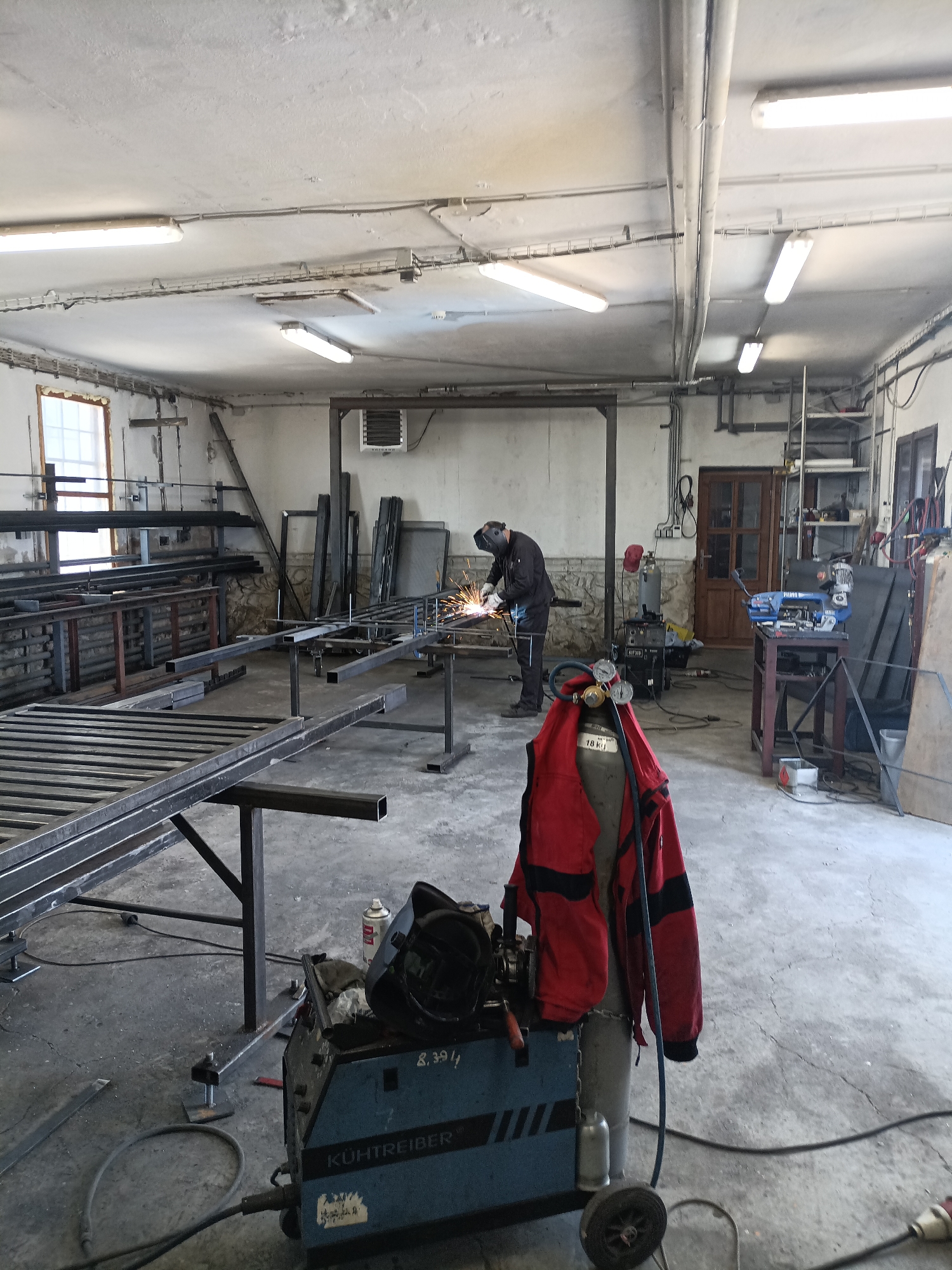 This image depicts an indoor workshop in Dalovy, Czechia, with a concrete floor and a high ceiling fitted with fluorescent lighting and exposed electrical conduits. The workshop is filled with metalwork, including long metal bars and frameworks arranged on benches and the floor. In the mid-ground, a person wearing dark work attire and a welding mask is actively engaged in welding, creating sparks. To the left foreground, a blue welding machine with the brand "KÜHTREIBER" is visible, alongside a red welding jacket draped over a gas cylinder. The environment suggests an active metal fabrication or repair facility.