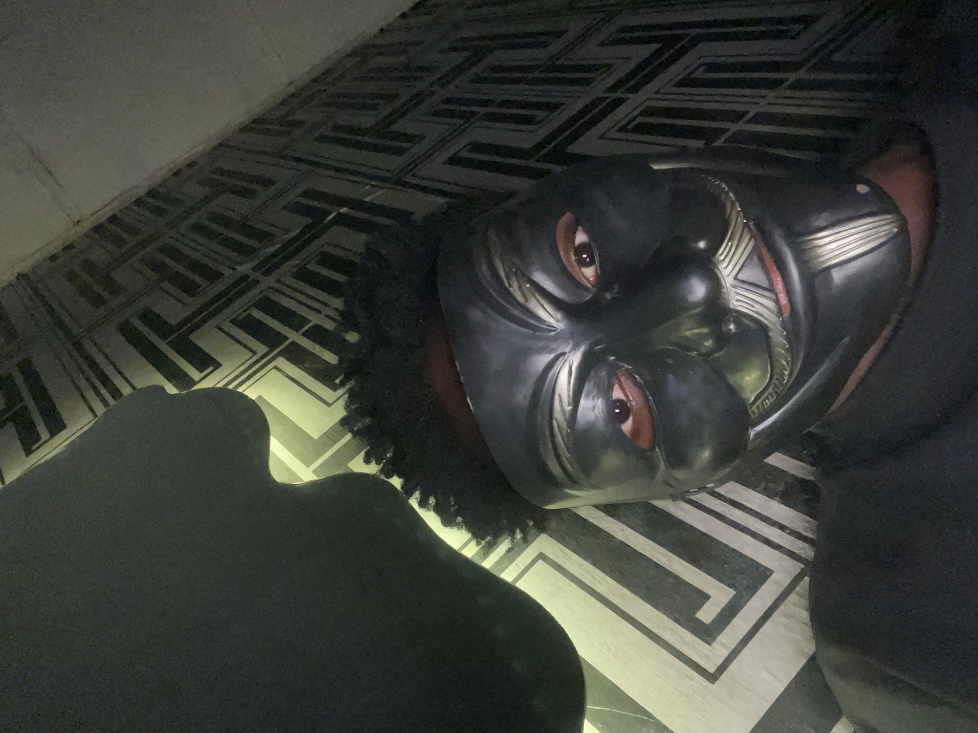 A person wearing a Black Panther mask is lying on a floor with a geometric pattern. The person has brown eyes and curly black hair visible around the edges of the mask. A dark object, possibly clothing or a blanket, is positioned next to the masked person. The location is not specified in the image, and there is no information to suggest it is in Lokoja, Nigeria.