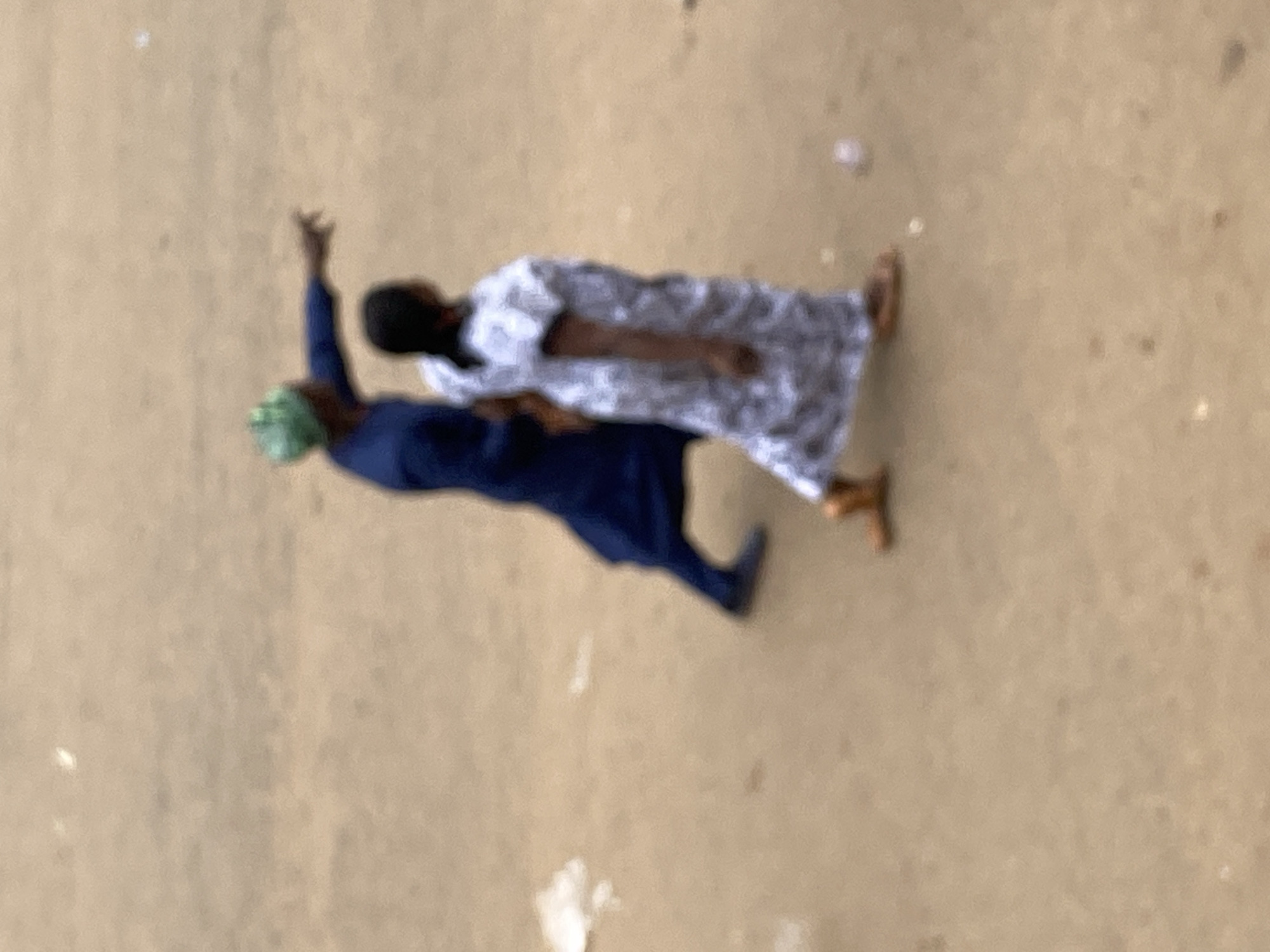 In Bukuru, Nigeria, two individuals are observed walking on a sandy surface. The person in front, facing away from the camera and to the right, is wearing a white and black patterned dress and brown sandals. The second individual, to the left and slightly behind, is wearing a dark blue garment and a green headscarf. This individual is gesturing with their right arm, pointing forward. The ground is a light brown, sandy terrain with some scattered debris, possibly including small white pieces of paper. The scene appears to be outdoors, possibly a street or an open area.