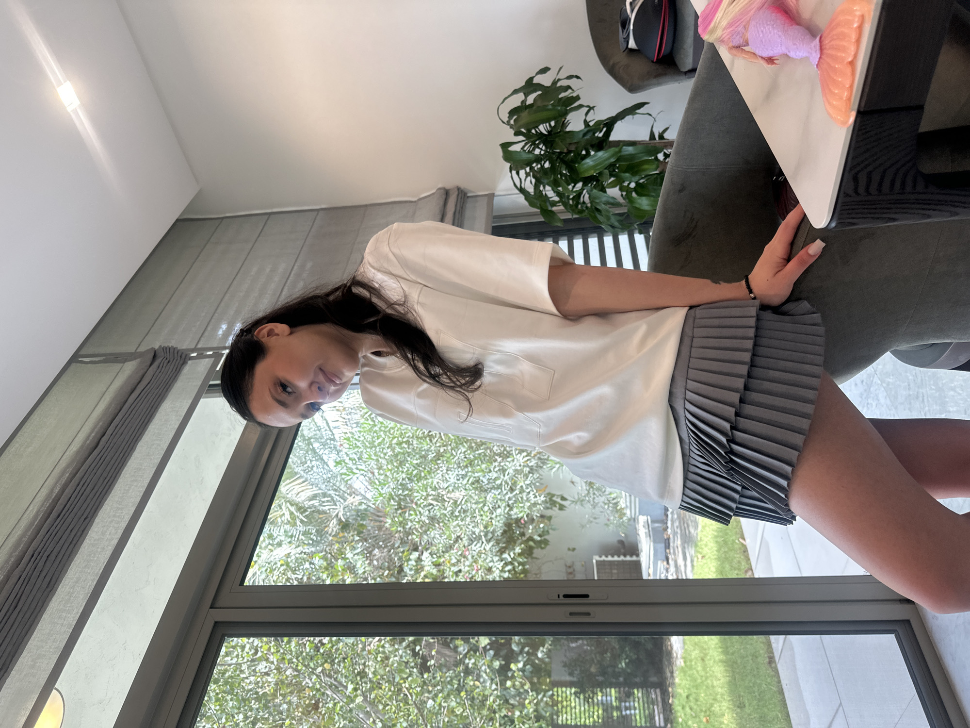 This image, originally oriented sideways, captures a young woman posing indoors next to a large glass window, looking towards the camera. She has long, dark hair and is dressed in a plain white short-sleeved t-shirt and a grey pleated mini-skirt. Her right hand, adorned with manicured nails and a dark beaded bracelet, rests on a dark grey upholstered chair or sofa.
The interior features white walls and ceiling, with a single recessed light fixture visible on the upper left wall. Grey blinds or curtains are partially drawn over the windows, which span a significant portion of the wall. On a white tabletop, a colorful pink and orange mermaid doll is visible.
Through the large windows, a vibrant outdoor scene unfolds, dominated by lush green trees and foliage, including what appears to be palm trees, suggesting a warm climate. A paved outdoor area can also be seen. The abundant natural light filling the room indicates it is daytime. The modern aesthetic of the indoor space, combined with the vibrant, green outdoor view, aligns with the context of a location like Dubai, United Arab Emirates. There is no visible text in the image.