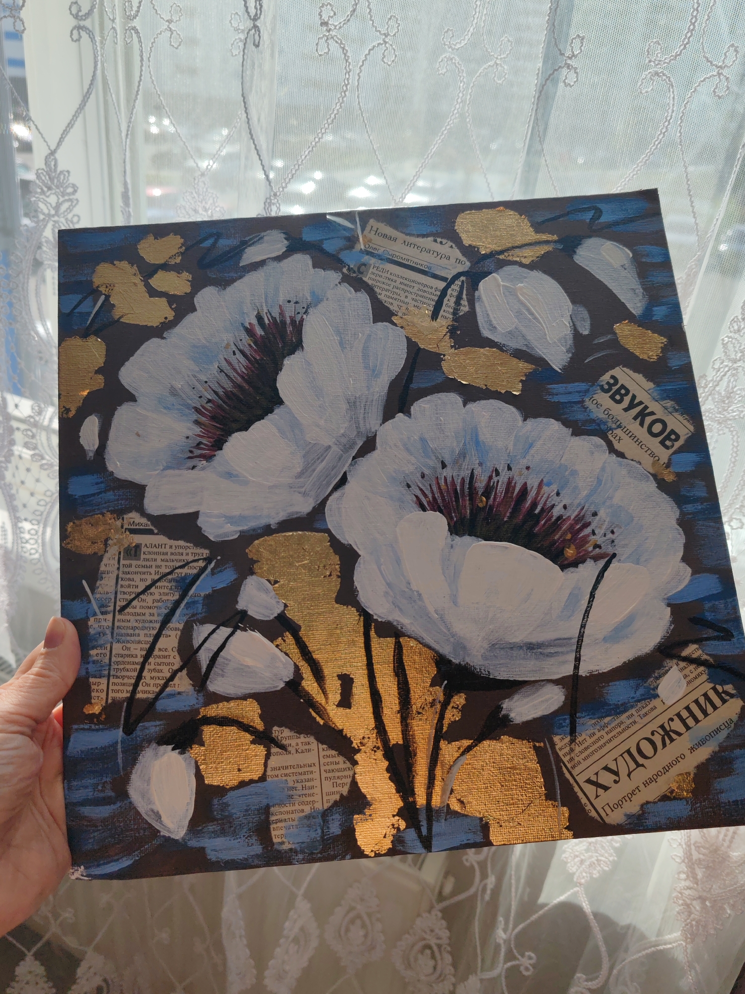 The image captures a mixed-media painting held by a person's hand, set against a bright window with white lace curtains. The artwork features two prominent white flowers with dark, possibly reddish-purple, centers, along with several smaller white buds. These floral elements are set against a dark background with textured blue horizontal strokes and scattered gold accents, which resemble abstract leaves or metallic flourishes. Thin black lines suggest stems or outlines.
Newspaper clippings, written in Russian, are integrated into the painting as collage elements. Visible text includes: "Новая литература по" (New literature on) and "Олег Сыромятников" in the upper left. In the upper right, a clipping reads "ЗВУКОВ" (of SOUNDS). In the lower right corner, a prominent piece of text states "ХУДОЖНИК" (ARTIST/PAINTER) followed by "Портрет народного живописца" (Portrait of a folk artist). Other discernible words from the various clippings include "Михай" (Mikhail), "АЛАНТ" (TALENT), "институт," "колекционеров" (collectors), and "литературы" (literature).
The scene is brightly lit by natural light, suggesting it is daytime. The soft, sheer curtains indicate an indoor setting, likely a home in Saint Petersburg, Russia. The visible hand suggests the painting is being displayed or presented. No specific emotional cues are discernible from the partial view of the person.