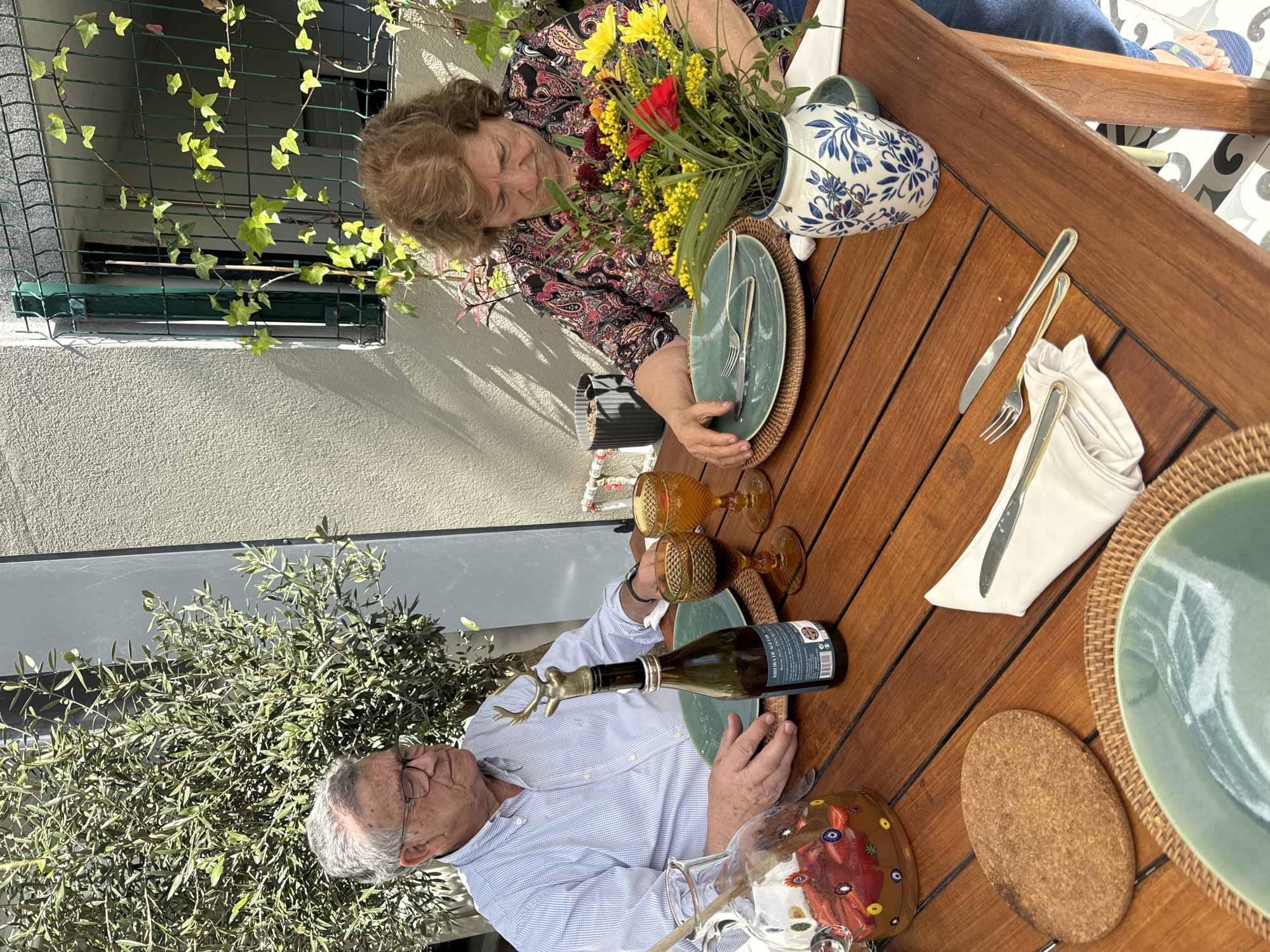 The image shows a wooden table set for a meal on a patio or outdoor dining area. Two people are present, an older woman and an older man. The woman is seated at the top of the frame, wearing a patterned blouse and looking down at the table setting. A vase of flowers is placed in front of her. The man is seated at the bottom of the frame, wearing a blue striped shirt. He is holding a bottle of wine with a decorative stopper. The table is set with green plates, woven placemats, cutlery, napkins, and amber-colored glasses. A colorful decorative carafe is also present. A window with ivy is visible in the background, as well as an olive tree. The location is likely Porto, Portugal, suggested by the architecture and outdoor setting. The scene depicts a couple preparing to share a meal outdoors.