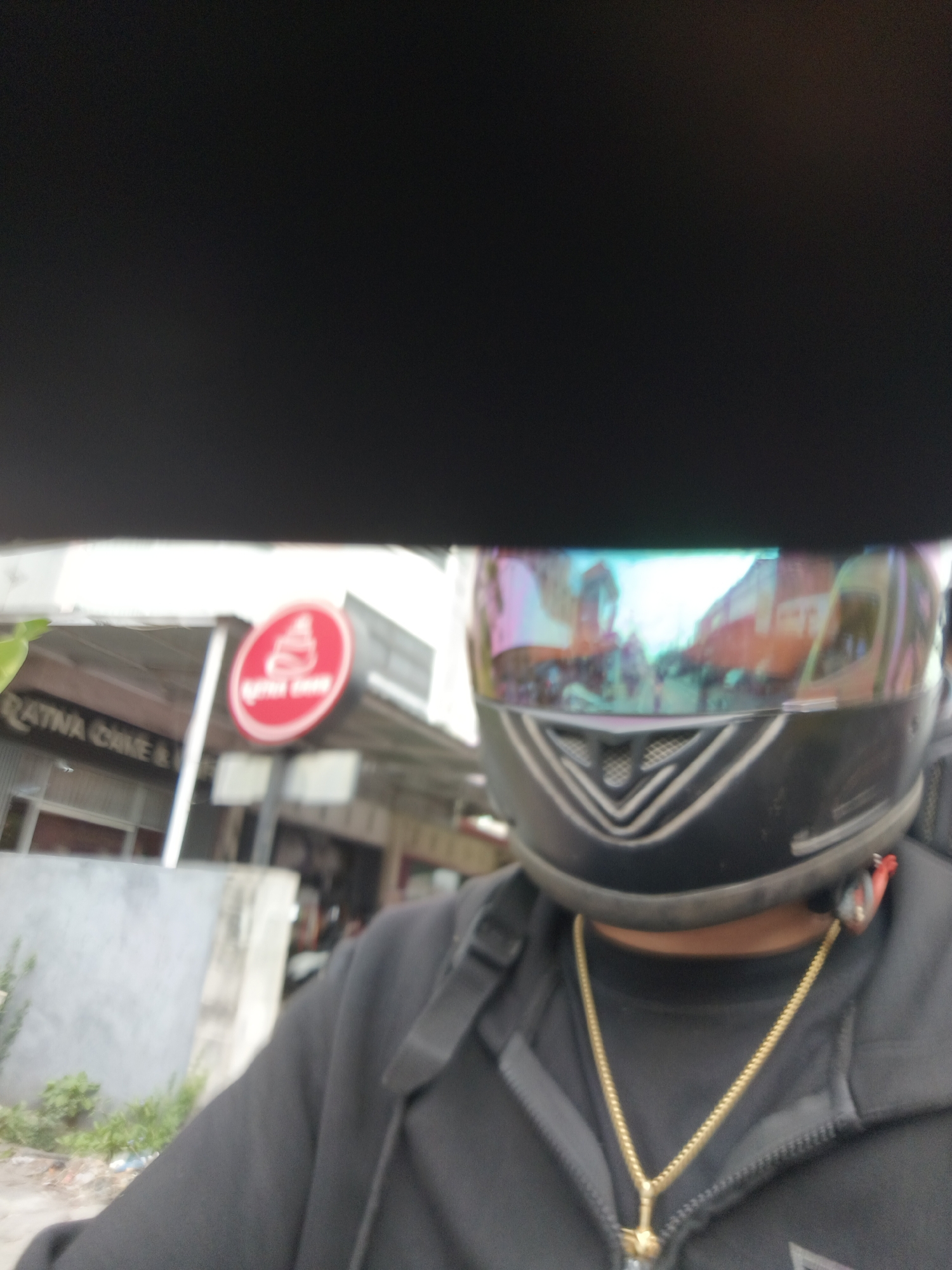 The image captures a first-person perspective of an individual wearing a black hoodie and a black motorcycle helmet with a multi-colored, reflective visor. The visor reflects a blurred image of a street scene, suggesting the subject is on a motorcycle. A gold chain necklace is visible around the subject's neck. In the background, a building with a sign that reads "RATNA CAKE & BAKE" is partially visible. To the right of the sign, a red circular logo with a white design is prominent. The environment appears to be an urban street in Denpasar, Indonesia, indicated by the architectural style and the likely presence of businesses like a bakery. The lighting suggests it is daytime, with a mix of sunlight and shadow.