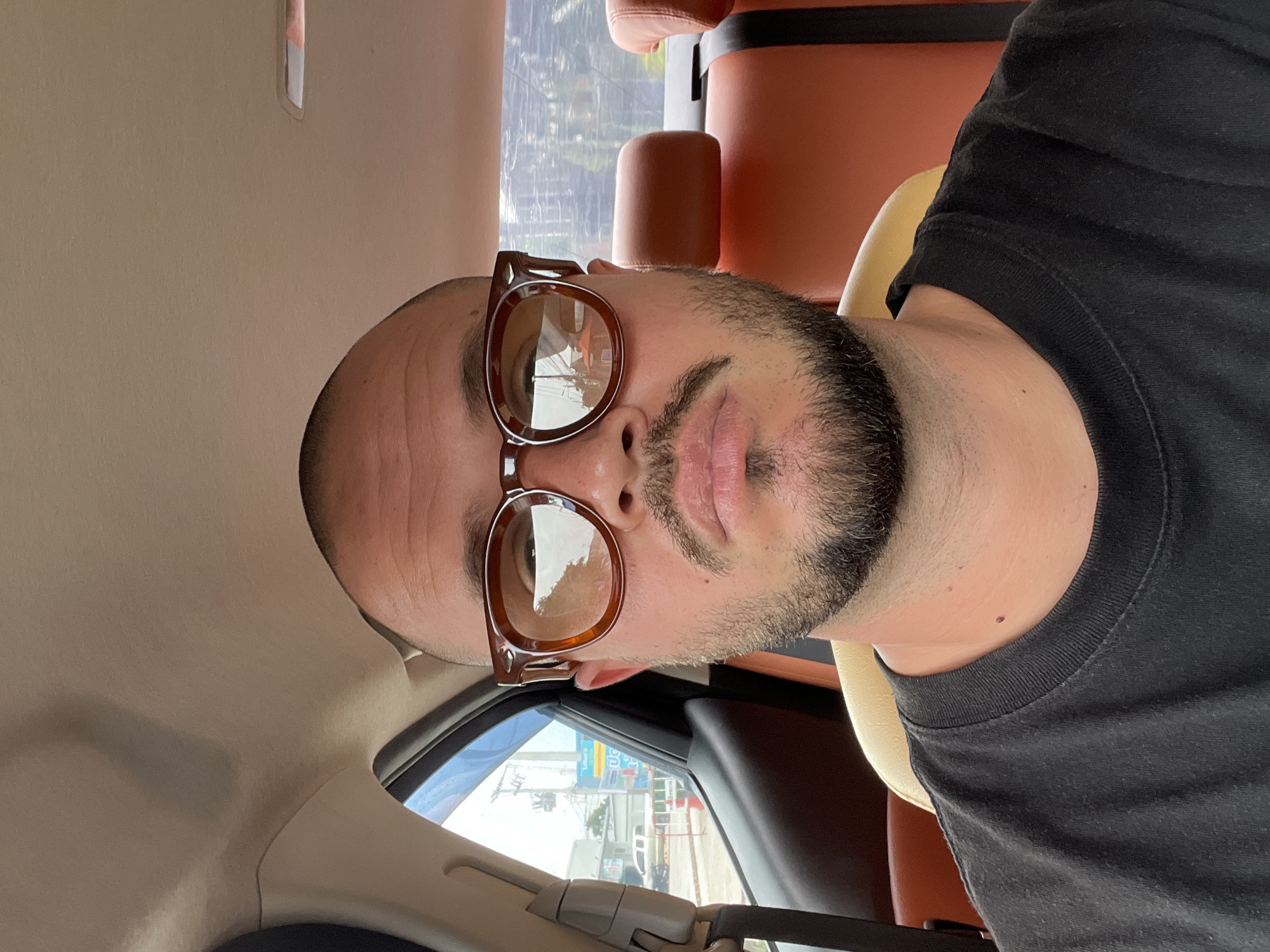 A man sits in the back seat of a car, wearing a black t-shirt and brown-rimmed glasses. He has a shaved head, a mustache, and a beard. The interior of the car has tan headrests and orange seats. Outside the car window, a street scene with buildings and a white truck is visible.