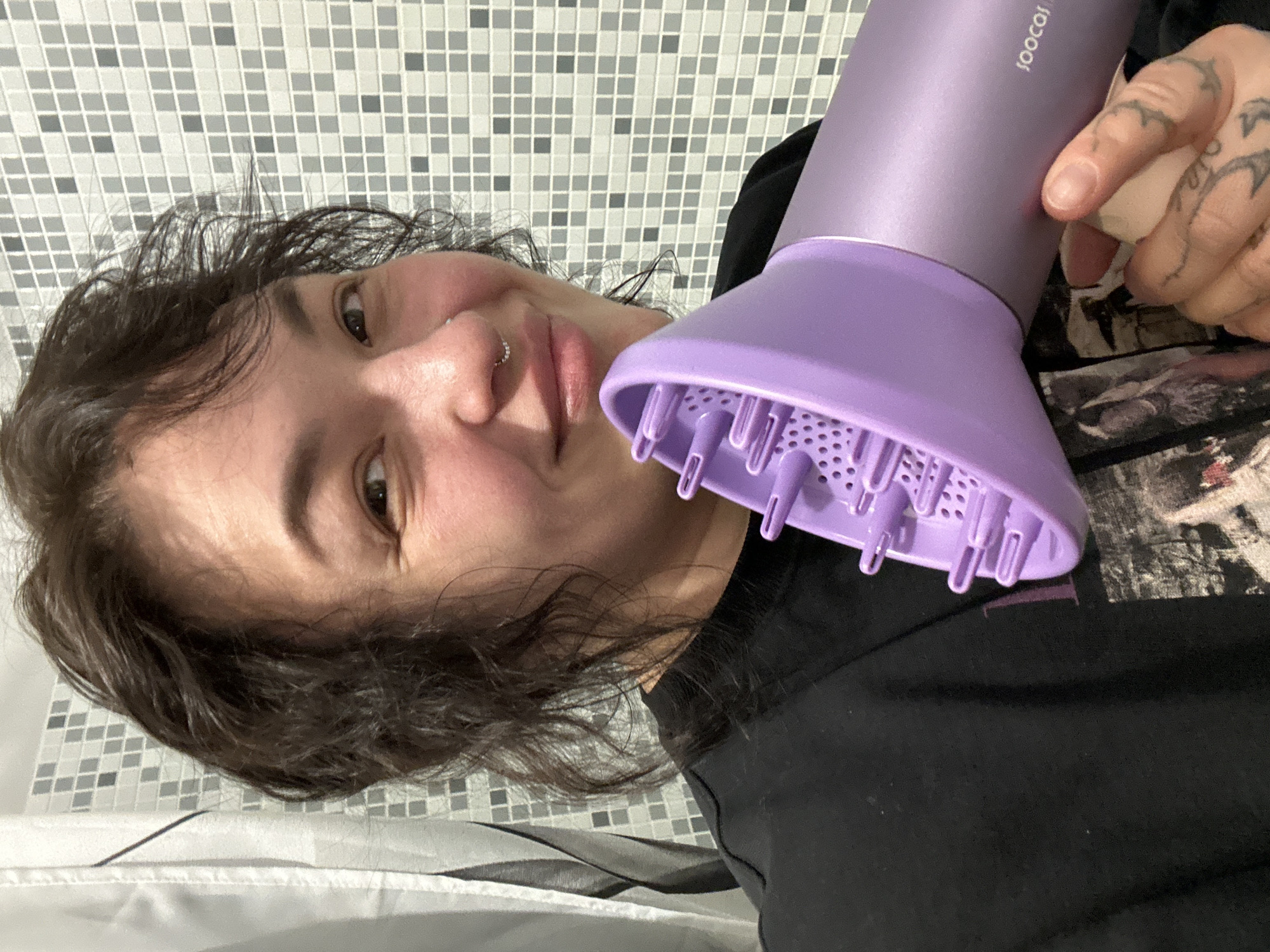 A person with dark hair and a nose piercing holds a purple hair dryer with a diffuser attachment close to their face. The person is wearing a black top. The background shows a tiled wall with a grey and white mosaic pattern. Part of a patterned garment lies on the floor. The brand "joocoi" is visible on the hair dryer. The location is likely indoors, possibly a bathroom. There's no information to suggest the image was taken in Chaikovsky, Russia.