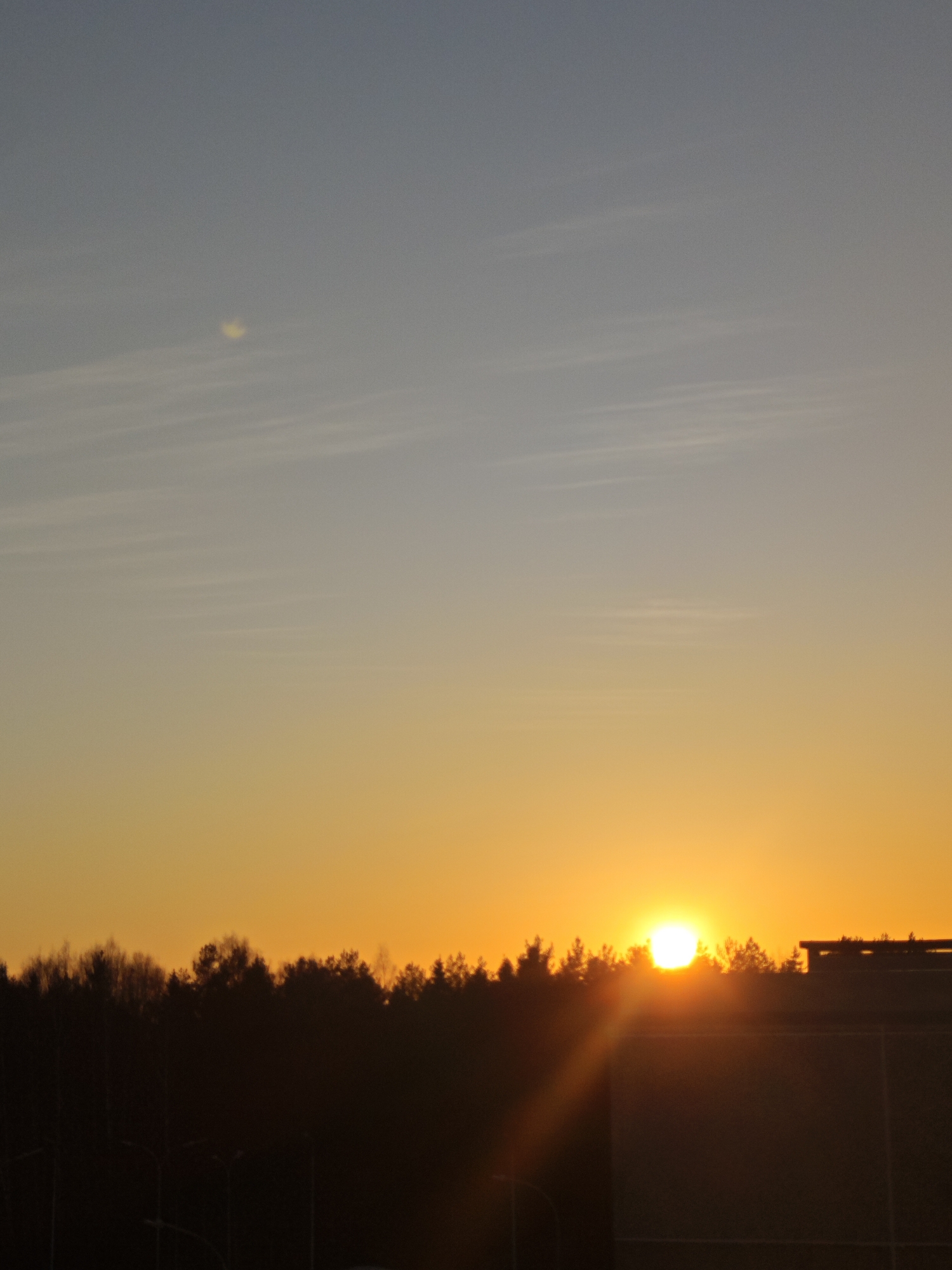 A wide shot depicting a sunset over a treeline and a building. The sky transitions from blue at the top to orange near the horizon. The sun is visible, partially obscured by the trees. The scene appears to be located in Markovskii, Russia, based on visual analysis.