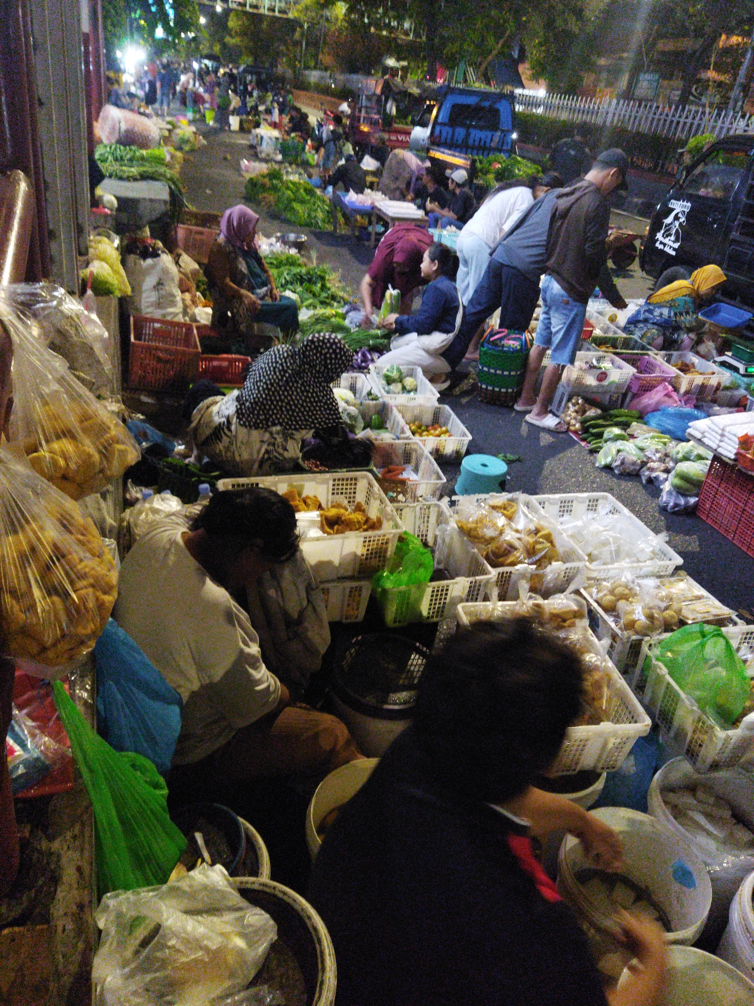 This is a medium-angle, eye-level, outdoor, nighttime photograph of a bustling street market in Semarang, Indonesia. The scene is filled with activity, with numerous people and stalls selling various goods, primarily produce. The market appears to be set up along a street, with vehicles, including a blue truck, present in the background.
In the foreground, on the left, several large, clear plastic bags filled with yellow, round items, possibly food items like snacks or fried snacks, are visible. To the right of these bags, a person wearing a light-colored long-sleeved shirt is bent over, their head down, with their hands partially obscured. They are near a white basket filled with more of the yellow food items.
Further into the frame, several people are engaged in market activities. A woman wearing a purple headscarf and a dark outfit is seated behind a display of green vegetables. Another person, wearing a black and white patterned head covering and clothing, is also seated among the produce. A man in a dark hoodie and blue shorts is standing and appears to be interacting with a woman who is wearing a yellow headscarf and patterned clothing.
The ground is paved, and the market is lit by overhead artificial lights, creating a vibrant and lively atmosphere. The stalls are laden with fresh produce, including leafy greens, and various packaged goods. The overall impression is of a busy, nighttime market scene characteristic of urban areas in Indonesia.