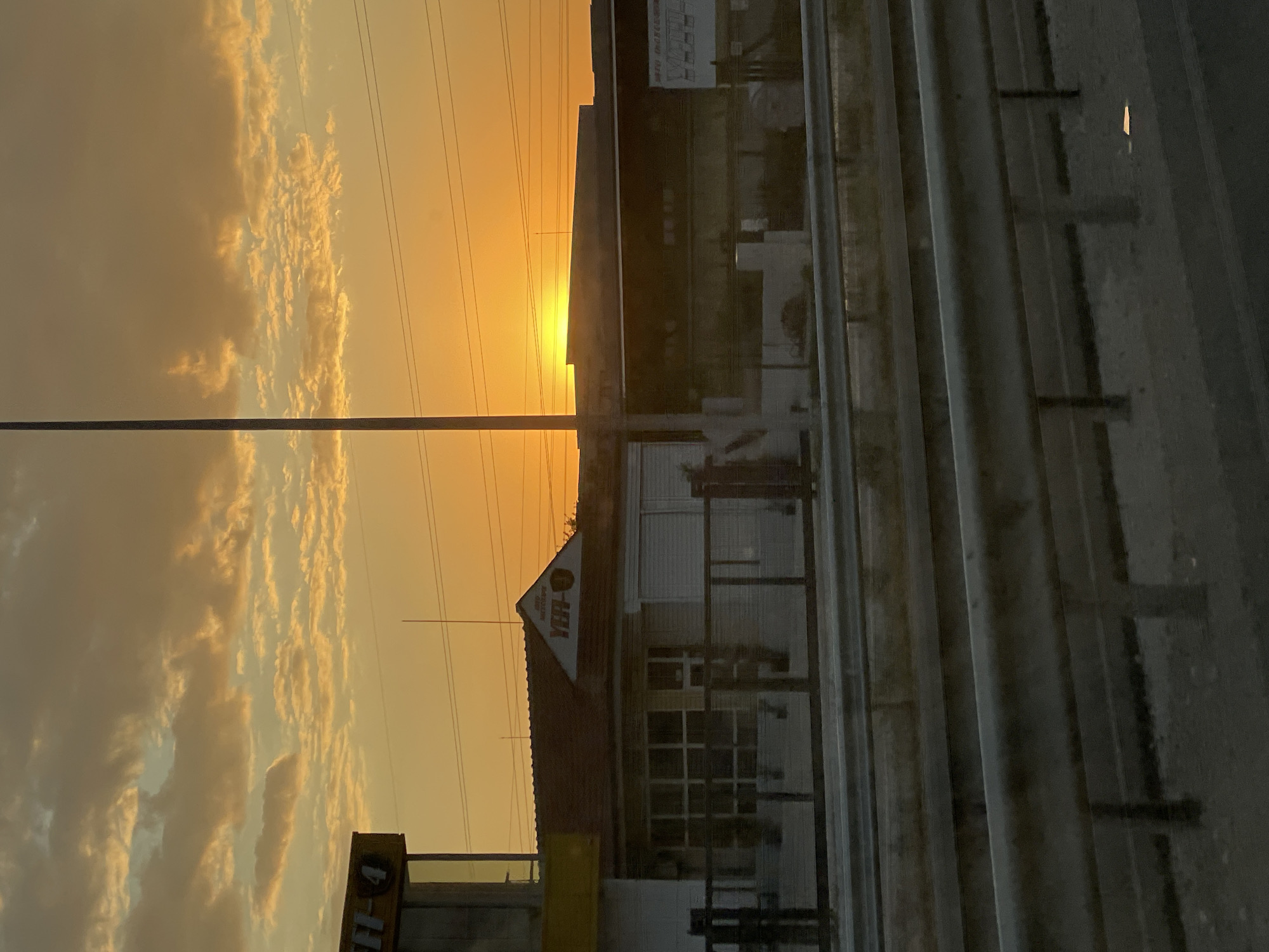 The image depicts a sunset scene with the sun low on the horizon, casting a warm orange glow across the sky. Scattered, textured clouds are visible against the bright sky. In the foreground, a highway guardrail runs horizontally across the frame, with the pavement and more guardrails visible beyond it. In the middle ground, a building with a red-tiled roof and white walls is situated. A sign with the text "VER-U" is visible on the building's gable. Another building with a dark facade and some signage is behind it, partially obscured. A tall pole stands prominently in the center of the image, extending from the ground towards the sky. The overall impression is one of an urban or semi-urban setting during twilight. This scene is located in Asenovgrad, Bulgaria.