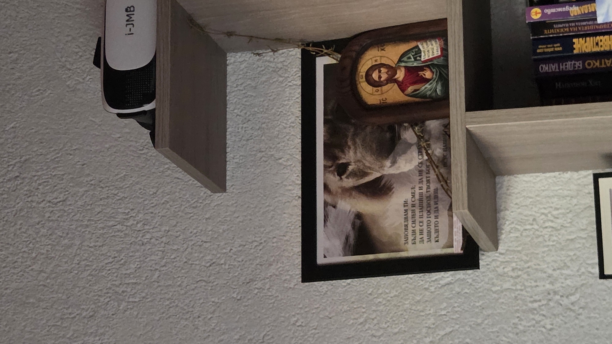 In an indoor scene, a VR headset is placed on a shelf. Below it, on another shelf, is a framed picture of a lion with text in Bulgarian, and an Orthodox icon of Jesus Christ. The wall behind the shelves has a textured finish. The shelves are part of a bookshelf containing several books. The overall environment appears to be a domestic setting.