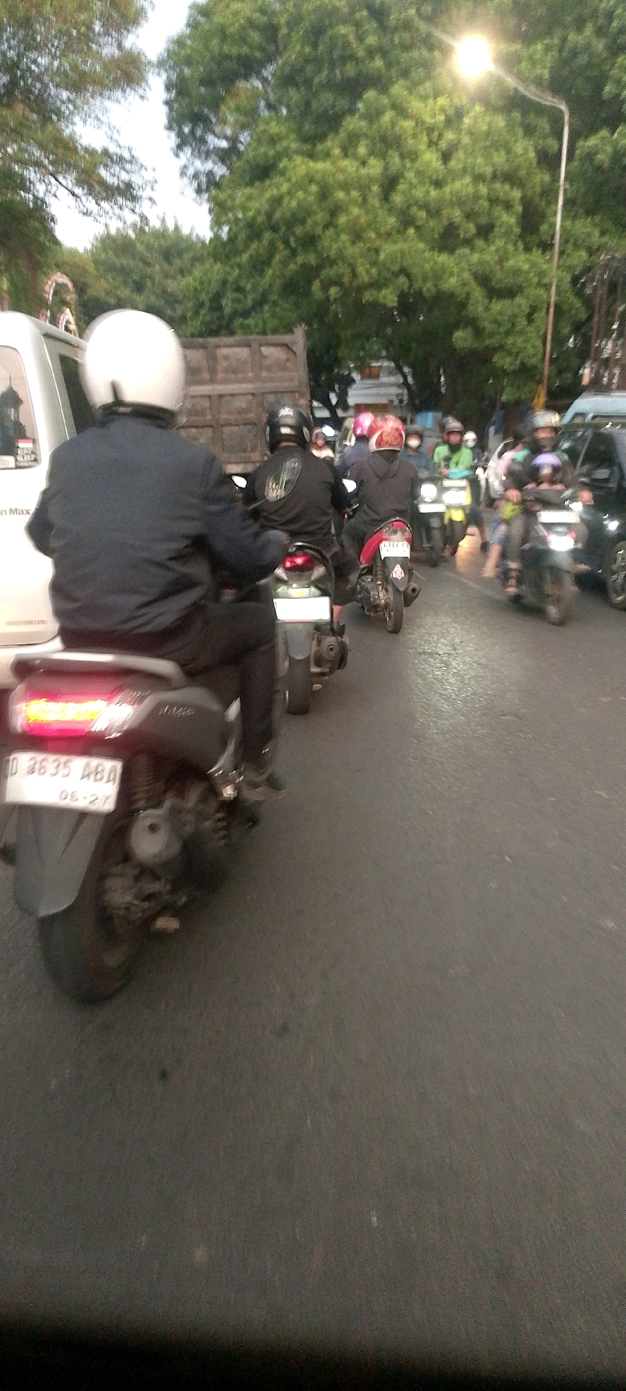 The image depicts a street scene in Bandung City, Indonesia, likely during daylight, with a procession of motorcycles and a truck. The foreground shows the rear of a motorcycle with a white license plate displaying "D 3635 ABA" and "06-27". The rider of this motorcycle is wearing a black jacket and pants, and a white helmet. Behind this motorcycle, other motorcycles are visible, some with riders wearing helmets. A large truck with a wooden bed is in the middle ground, and further back, more motorcycles and vehicles are on the road. Trees with green foliage line the side of the road, and a street light is illuminated, suggesting overcast conditions or dusk. The road surface is asphalt. The overall impression is of moderate traffic congestion on a city street.