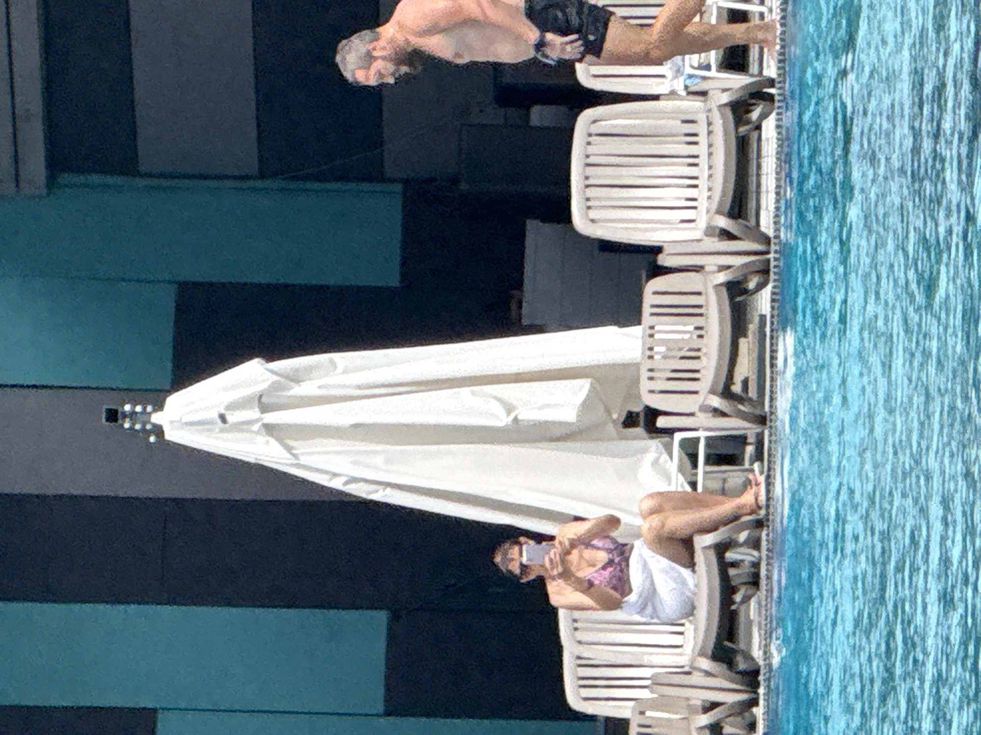 A man and a woman are by a swimming pool, which is located in the city of Массандровский поселковый совет, Ukraine. The woman, wearing a pink bikini and a white towel, is sitting on a lounge chair, looking at her phone. She has sunglasses on. In front of her is a large white umbrella. A man, wearing black swim trunks, is walking to the right of the frame, away from the pool. He has a beard and is looking to his left. The poolside has multiple empty lounge chairs. The background is a dark, tiled wall with vertical stripes of dark blue and teal.