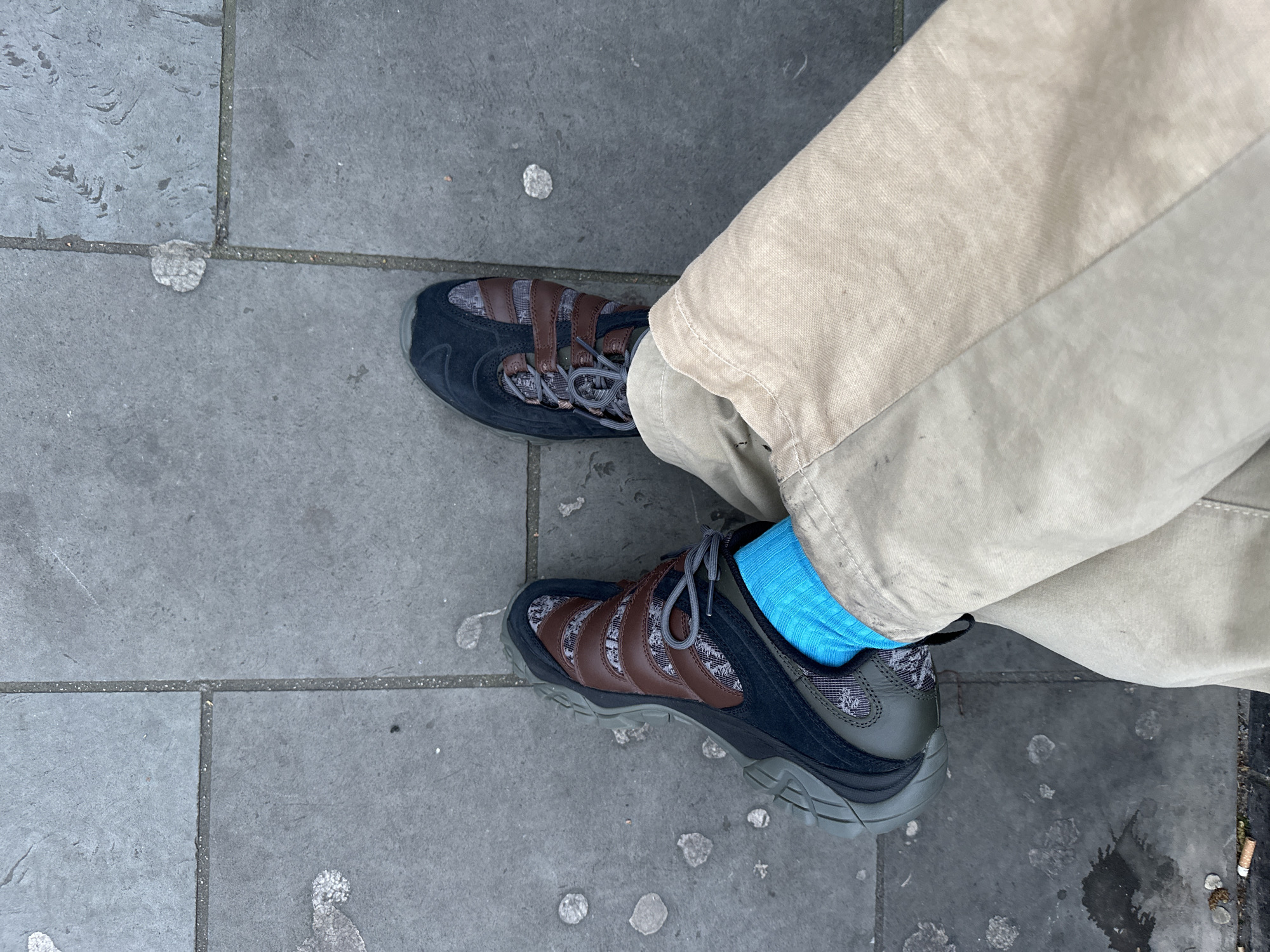 The image shows a pair of feet on a grey, paved surface, likely a sidewalk. The person is wearing beige trousers, a bright blue sock on the right foot, and brown and black athletic shoes with grey laces. The pavement is marked with what appears to be discarded chewing gum. In the lower right corner of the frame, cigarette butts are visible. Considering the prevalence of paved sidewalks, chewing gum residue, and cigarette litter, it is plausible that this photo was taken in an urban setting such as the city of London, United Kingdom. There is no specific feature of the location to confirm if it is in London.