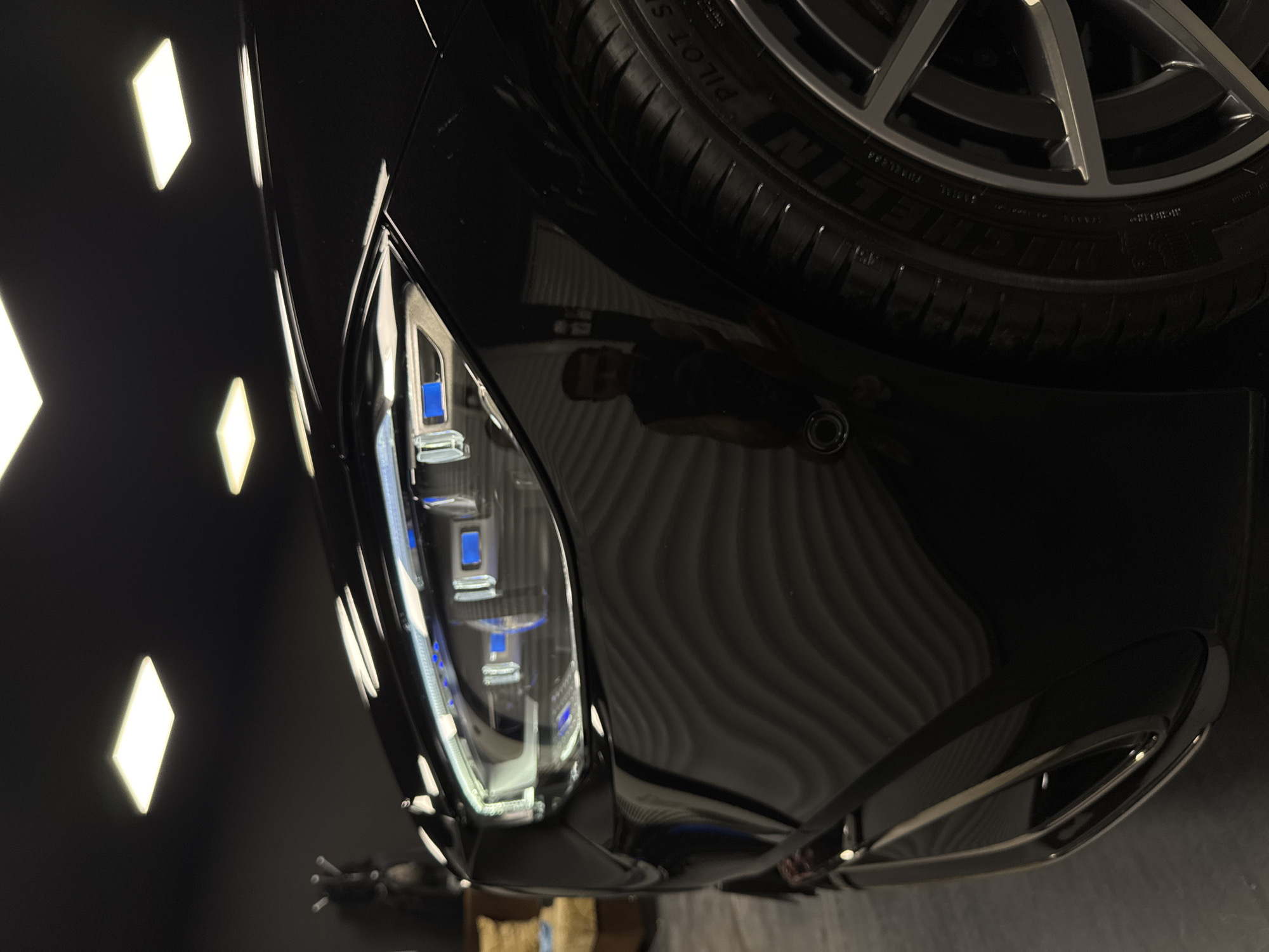 The image shows a close-up, low-angle view of the front passenger side of a black car, likely in a professional detailing or auto body shop in Ruse, Bulgaria. The car's sleek, glossy surface reflects the overhead fluorescent lighting panels on the ceiling and the outline of a person taking the photograph. The car features a complex headlight assembly with intricate LED elements glowing blue. The tire is a Michelin Pilot Sport, and the rim is a multi-spoke alloy wheel. The background is dark, with the ribbed texture of a garage door visible on the right. The overall impression is one of a high-end vehicle in a meticulously maintained environment.