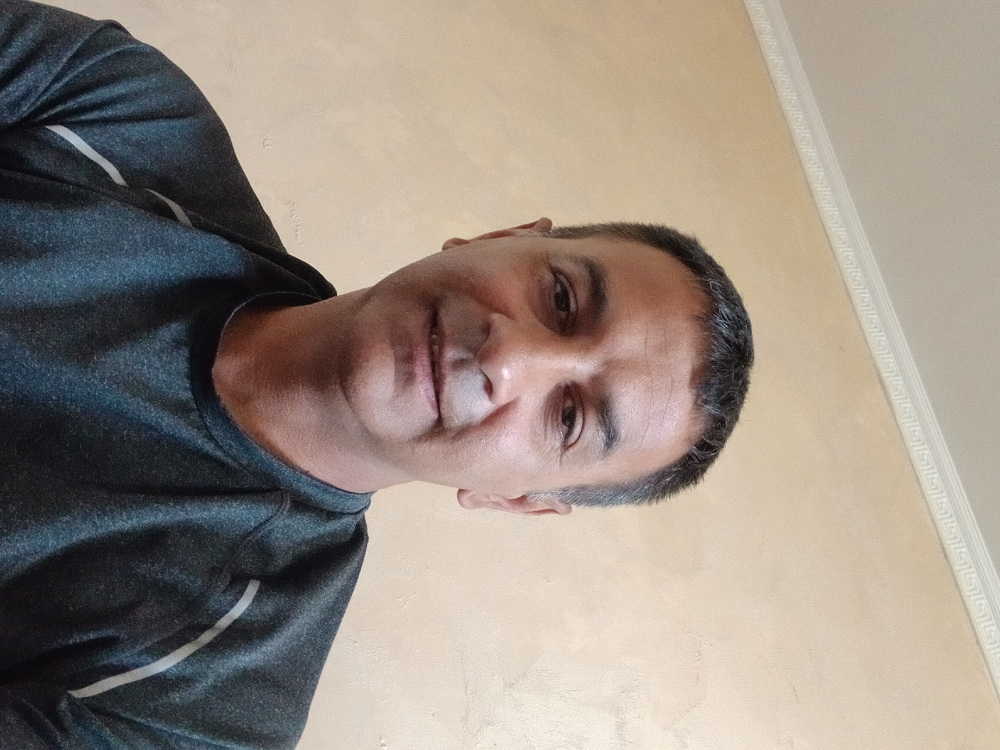A close-up, tilted portrait shows a middle-aged man with short, dark hair graying at the temples. He has brown eyes and a slight smile. He's wearing a dark gray shirt with a light gray stripe. The background is a textured light beige wall, with a decorative white molding along the top edge. The location is specified as Topolovgrad, Bulgaria.