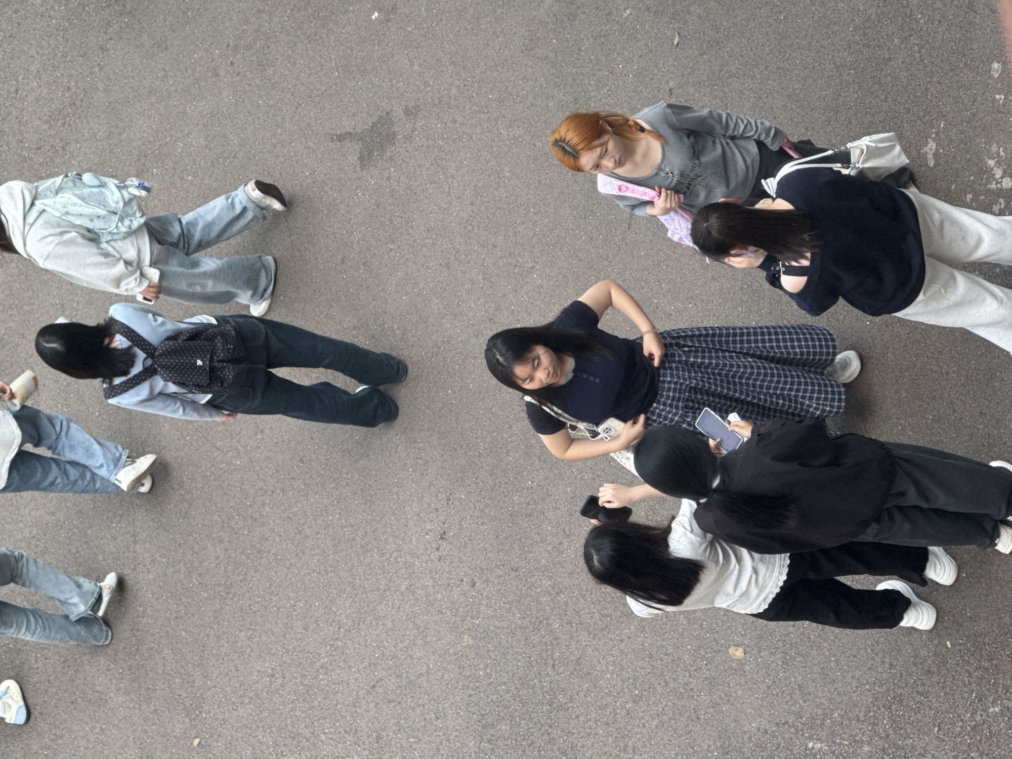 The image is an aerial view of a group of young women on an asphalt surface. It is possibly in Jiangjin, China but the location is not definitively known.
The group is made up of approximately 10 individuals. All the individuals are women. Their ages appear to range from late teens to early twenties. They are wearing casual clothing and white sneakers. Some of the women are standing while some are sitting. Several of the women are looking at their phones. One of the women has red/orange hair.
The setting appears to be a public outdoor space, possibly a plaza or large walkway. The asphalt surface is gray and shows signs of wear and minor discoloration. There are no prominent landmarks or features visible in the frame other than the ground surface and the people.