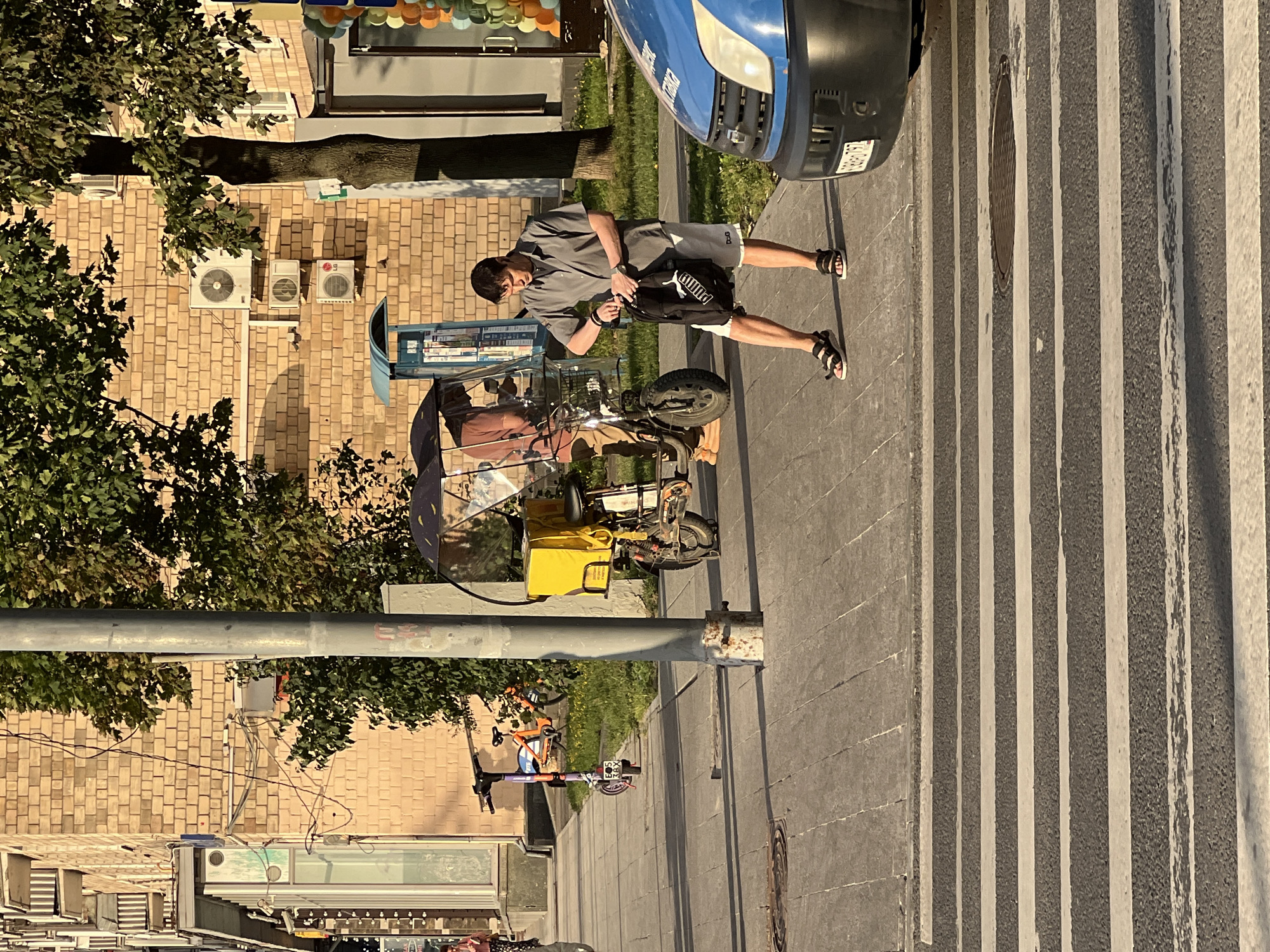 The image depicts a street scene in Moscow, Russia, on a sunny day. In the foreground, a pedestrian crossing is visible. A man wearing a gray short-sleeved shirt and white shorts with black sandals stands on the sidewalk, holding a black backpack with a white Puma logo. He appears to be adjusting something inside the backpack. To his left, a delivery scooter with a yellow insulated box is parked, covered by a clear plastic dome. Behind the scooter, another scooter with an orange frame is parked. In the background, a brick building with multiple windows and balconies lines the street. Several air conditioning units are mounted on the building's exterior. A tall metal pole stands prominently in the mid-ground. A blue vehicle is partially visible on the right side of the frame, positioned near the pedestrian crossing. The overall environment suggests an urban setting with commercial and residential elements.