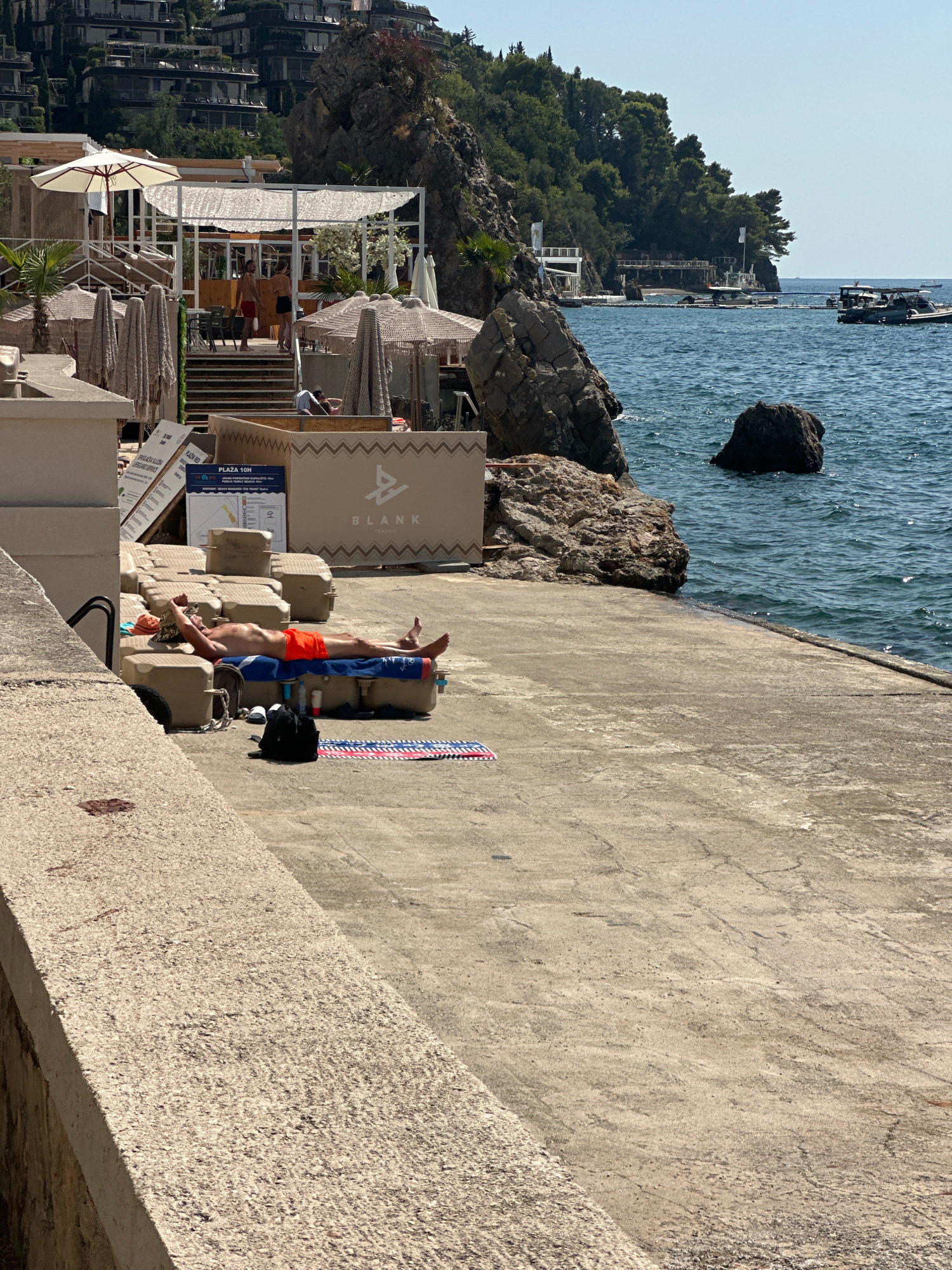 The image depicts a man sunbathing on a concrete pier by the sea in Budva, Montenegro. The man is lying on his back on a cushioned lounger, wearing orange swim trunks. Next to him are several flotation devices, a backpack, and a towel. In the background, there is a beach club with white canopies and umbrellas, and a rocky cliff face covered in lush green vegetation. Several boats are visible in the calm blue water, and a few buildings are nestled on the hillside in the distance. The overall scene is one of a relaxed summer day at a coastal resort.