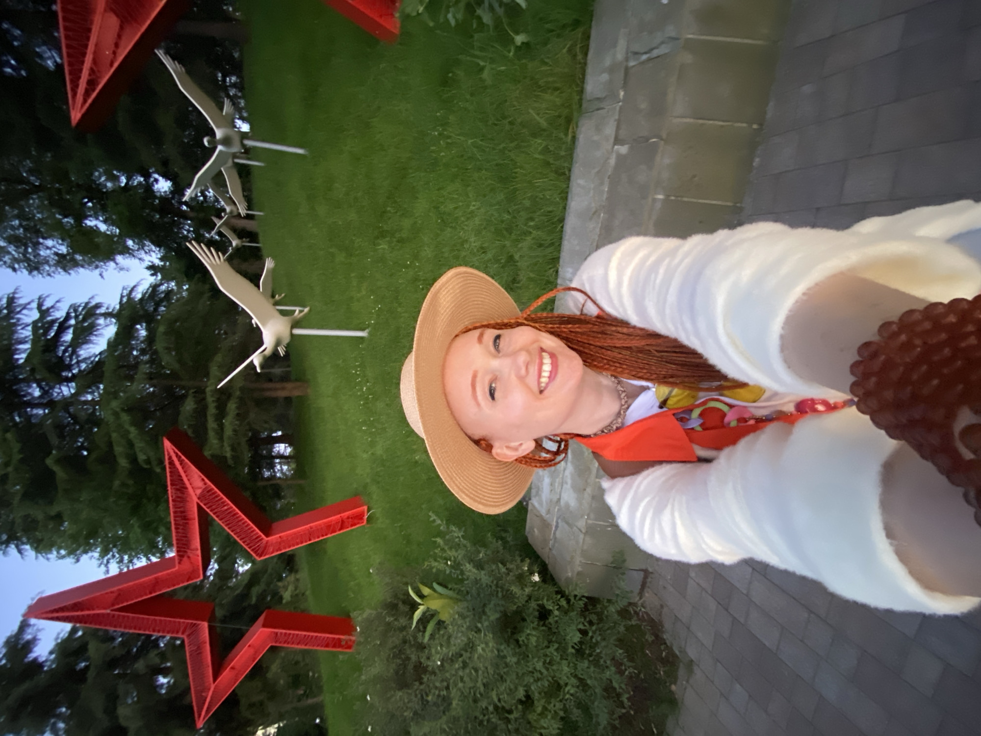 A young woman with reddish-brown braided hair and a wide, joyful smile is captured in a selfie taken in an outdoor park in Sochi, Russia. She wears a light-colored straw hat, a white fluffy long-sleeved top or jacket, and a colorful scarf or patterned top underneath. A beaded necklace and a large red beaded bracelet on her right wrist add to her vibrant appearance.
The scene, illuminated by the soft light of twilight or early evening, features a lush green lawn and numerous trees, including dense evergreen foliage. The park is adorned with significant sculptures: two large, red, multi-faceted star structures are visible, one partially in the foreground and another further back. Several light-colored bird sculptures, resembling cranes, are mounted on poles, creating the impression of birds in flight among the trees. These elements, particularly the red stars and cranes, often carry patriotic or memorial significance in Russian culture. A gray paved pathway or sidewalk runs along the right side of the image. The woman's cheerful expression conveys a sense of happiness and enjoyment in this scenic and culturally resonant location.