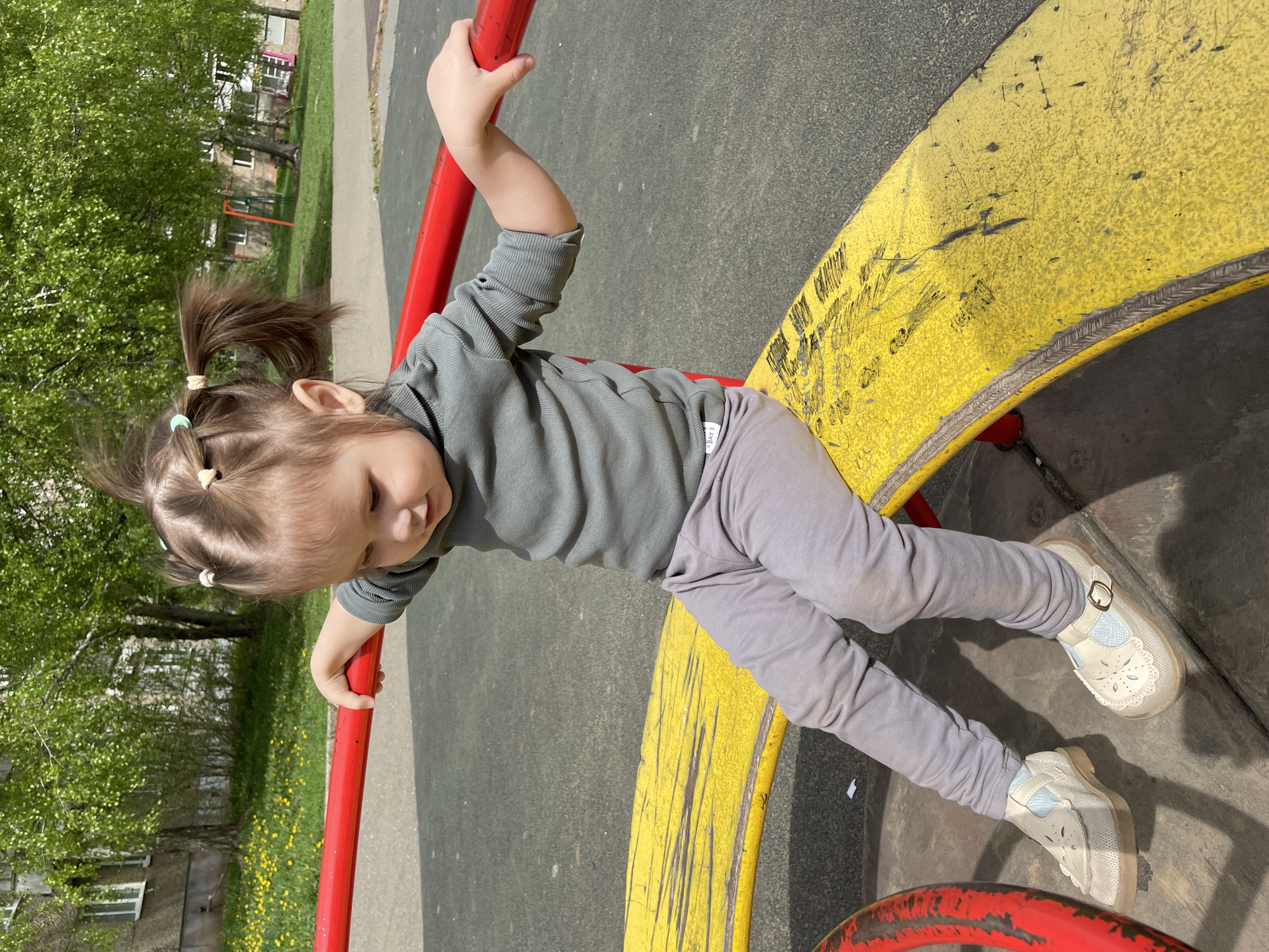 A young child is on playground equipment. She is gripping onto a red metal bar with both hands. She is wearing a grey shirt, grey pants, and light-colored shoes. The playground equipment has yellow paint on it. The playground is located in Izhevsk, Russia. There are trees and buildings in the background.