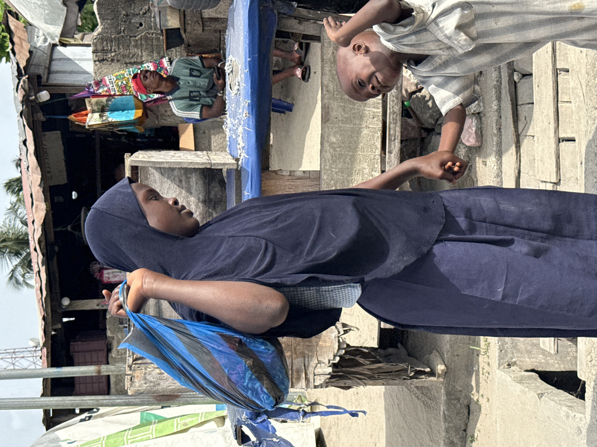 The image shows a woman in Ado, Nigeria, wearing a dark blue hijab and carrying a blue and black plastic bag. She appears to be interacting with a young boy in a striped shirt who is reaching out to her. In the background, a man sits under a blue covering, and there are buildings of modest construction typical of the city of Ado. The environment appears to be somewhat impoverished, with evidence of concrete construction and dilapidated structures.