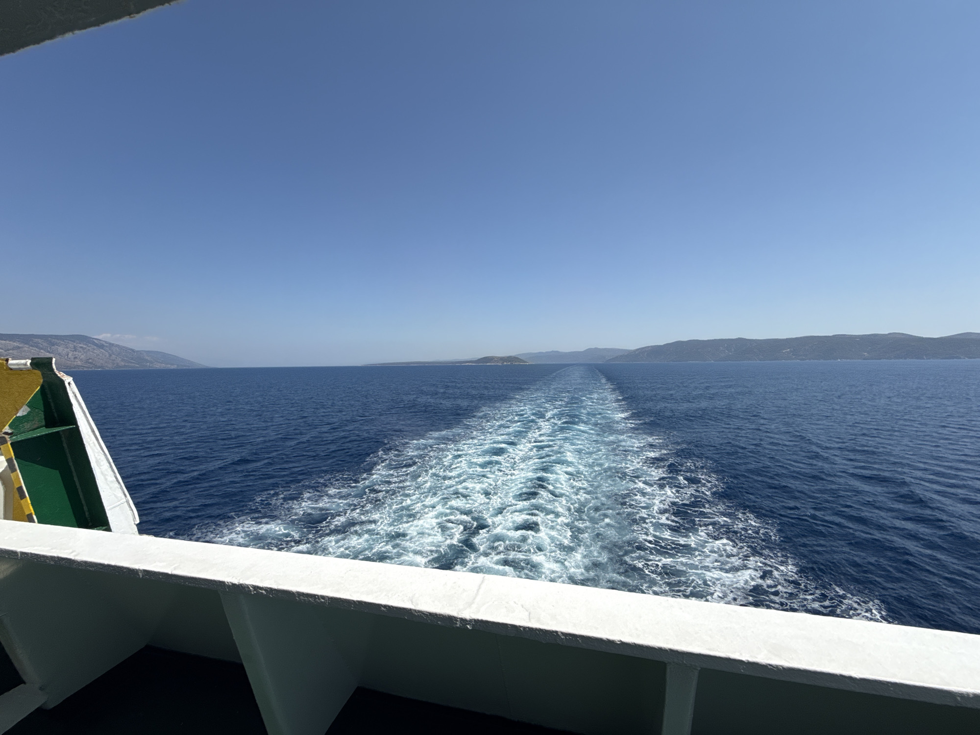 The image captures a view from the deck of a ferry boat traveling on the Adriatic Sea, likely near the town of Nerežišća, Croatia. The foreground is dominated by the white railing of the ferry, with a portion of its green and yellow hull visible on the left. The ferry is creating a prominent white wake as it moves through the deep blue water. In the distance, the hazy outlines of the island of Brač, where Nerežišća is located, can be seen along the horizon under a clear, bright blue sky. There are no people or animals visible in the frame. The scene is one of a journey at sea on a sunny day.