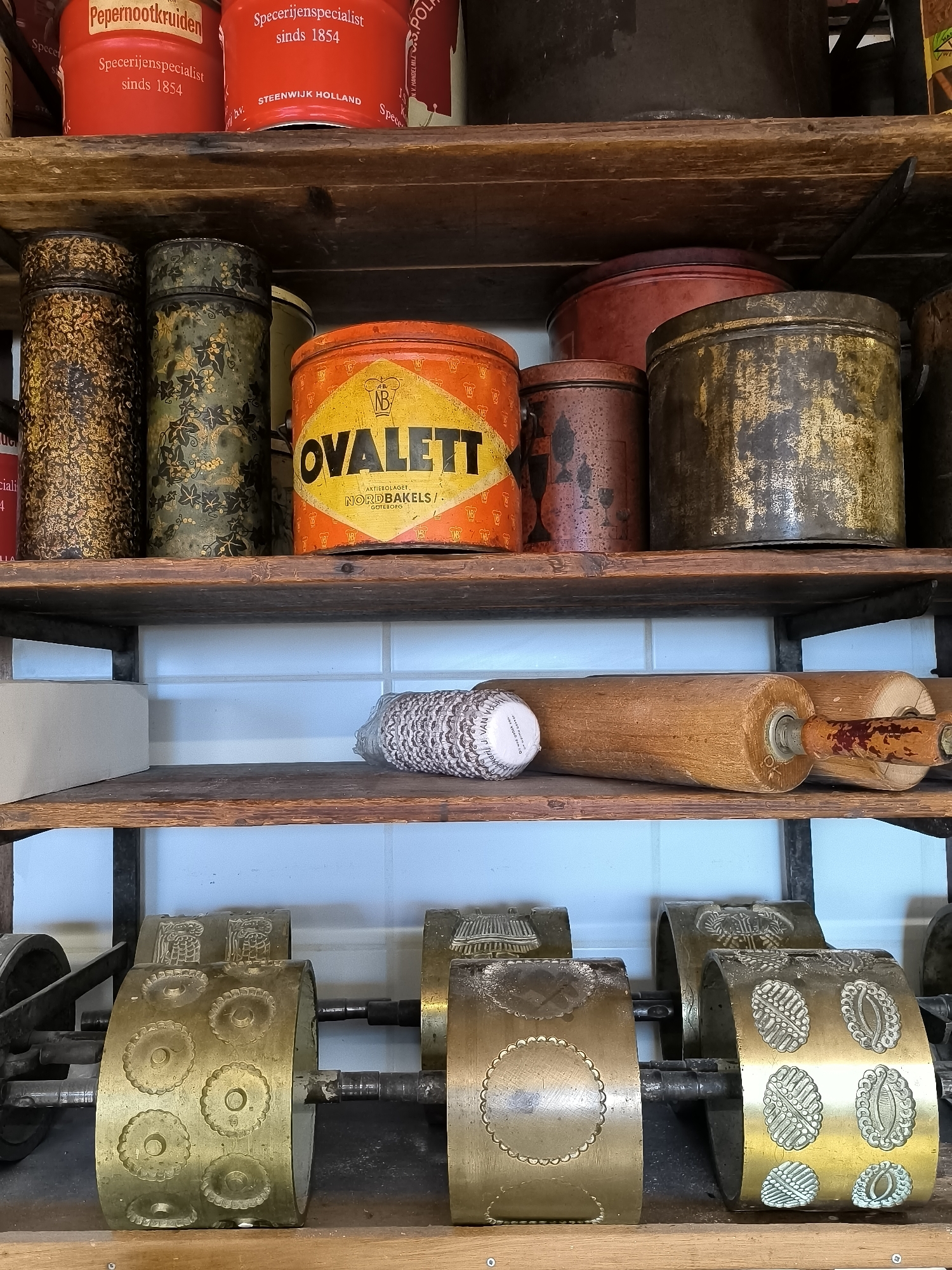 The image shows a three-tiered wooden shelf filled with an assortment of vintage baking supplies. On the top shelf, there are multiple cylindrical tins, varying in size, color, and design. The tins include red ones labeled "Pepernootkruiden" and "Specerijenspecialist sinds 1854," as well as decorative tins and a yellow and orange tin labeled "OVALETT" from Aktiebolaget Nordbakels in Goteborg. The middle shelf features a patterned paper cupcake liner and two wooden rolling pins, one with red paint on the handle. On the bottom shelf, there is a collection of brass cookie cutter rollers with various patterned designs. The backdrop behind the shelves consists of light blue tiled walls. This image was taken in Medemblik, Netherlands.