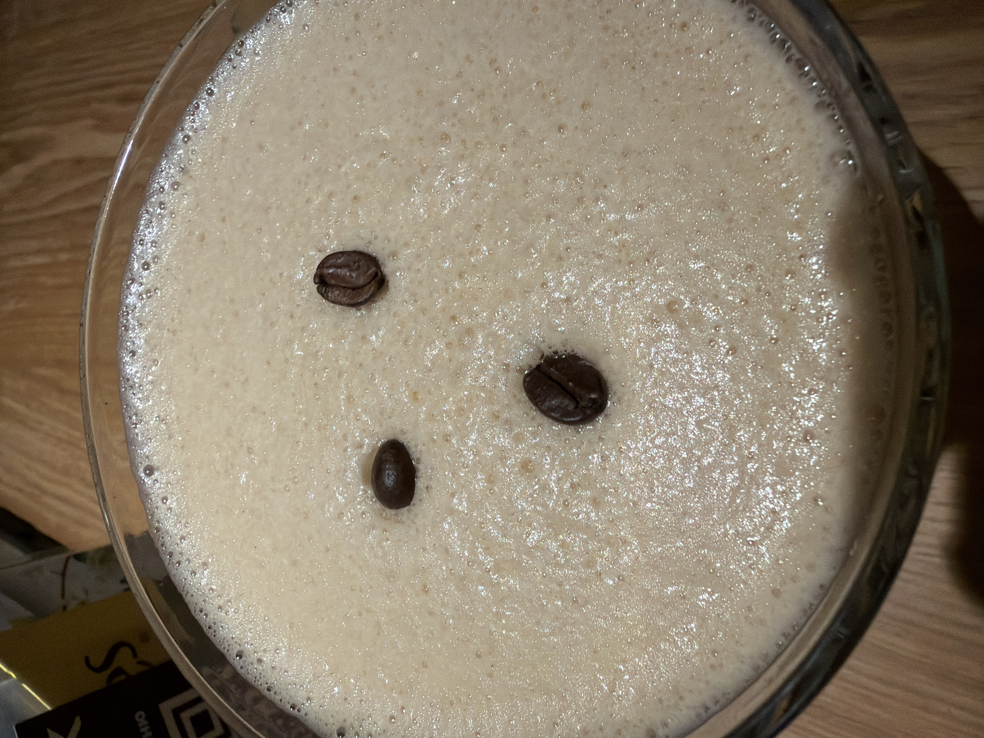 The image shows a close-up, overhead view of a cocktail in a stemmed glass, likely an espresso martini, decorated with three coffee beans arranged asymmetrically on top of the foamy surface. The drink itself is a pale, creamy brown color with numerous small bubbles creating a textured surface. The glass is clear and the liquid fills it nearly to the rim. The background is a blurry mix of browns and yellows suggesting a wooden table surface and possibly other objects nearby. There is no indication of the location being Varna, Bulgaria.