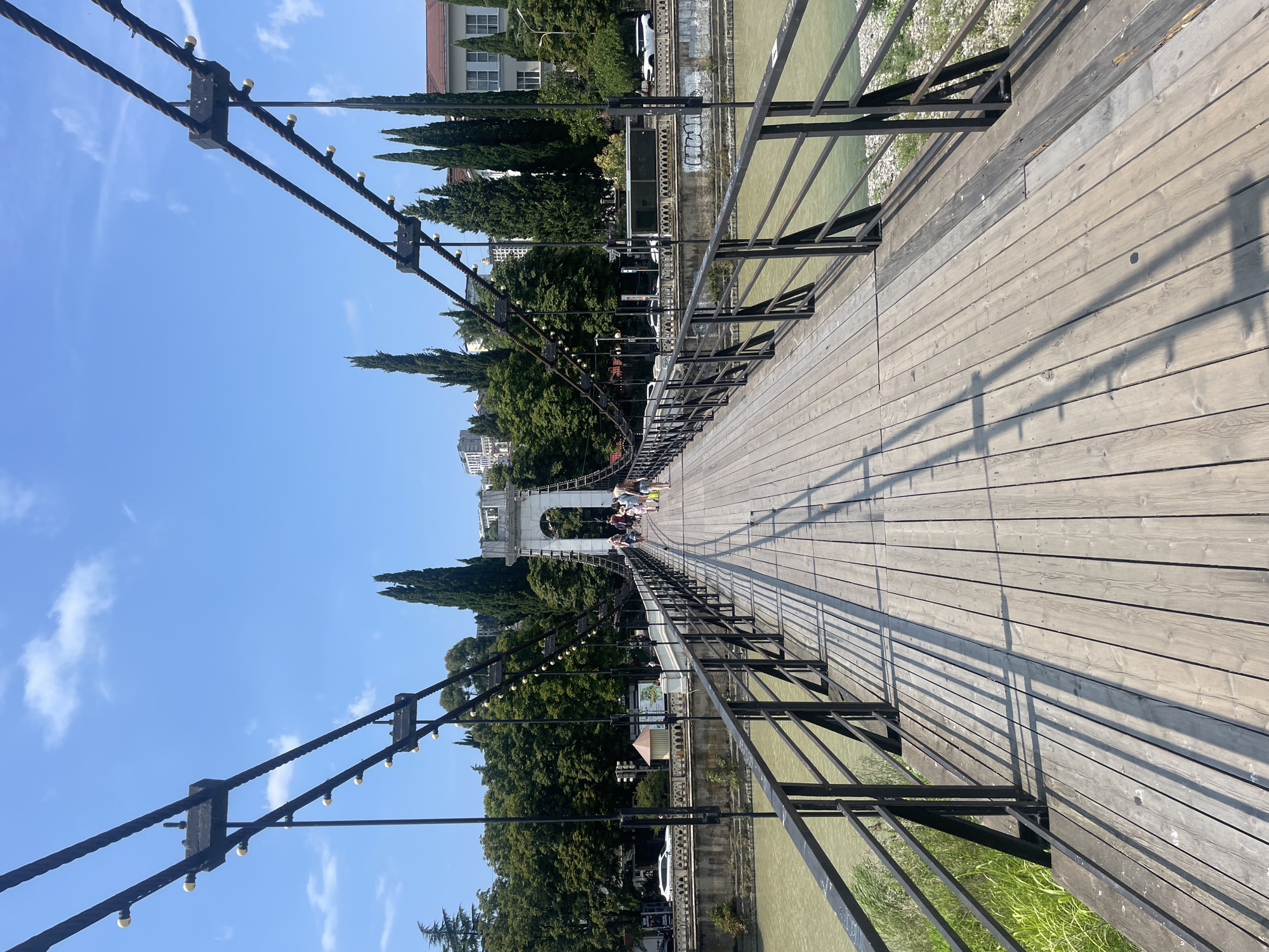 The image shows the pedestrian walkway of a suspension bridge in Sochi, Russia. The walkway is made of wooden planks with black metal railings on either side. Several people are walking across the bridge, some appearing to be tourists. The bridge spans a wide, murky river. In the background, there are green trees, buildings, and a clear blue sky with scattered clouds. The bridge's suspension cables are visible, as are decorative lights strung along them. The scene appears to be a sunny day, casting distinct shadows from the railings onto the wooden walkway.