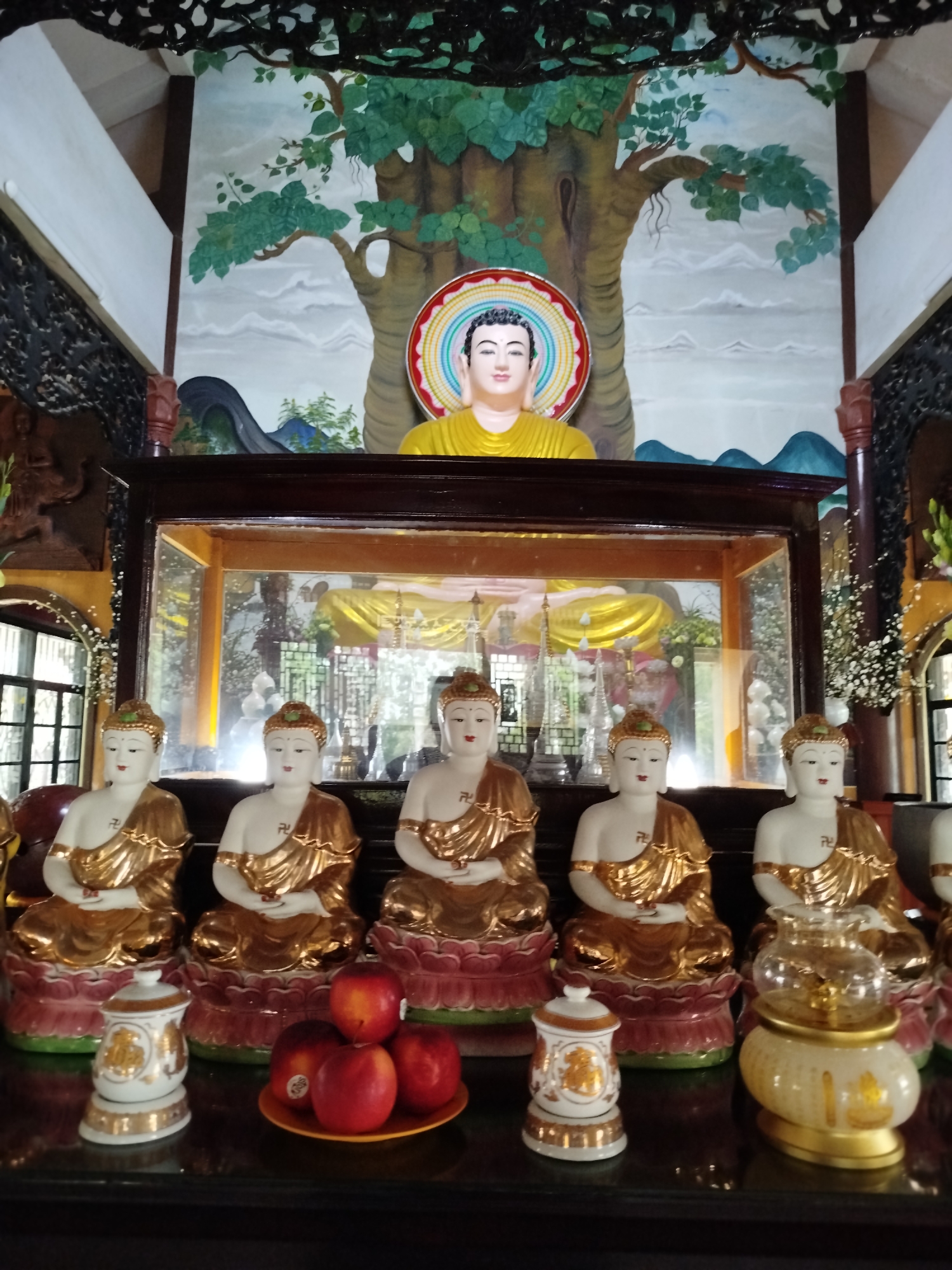 Captured in Xuân Trường, Đà Lạt, Vietnam, the image showcases a Buddhist altar. The altar is centered with five seated Buddha statues, each draped in golden robes, resting on lotus-shaped bases. Below them is a plate of red apples and decorative ceramic pots. Above, a glass-enclosed space contains a larger Buddha statue. A mural depicting a tree and landscape hangs behind the altar. Carved wood details the surrounding architectural elements.