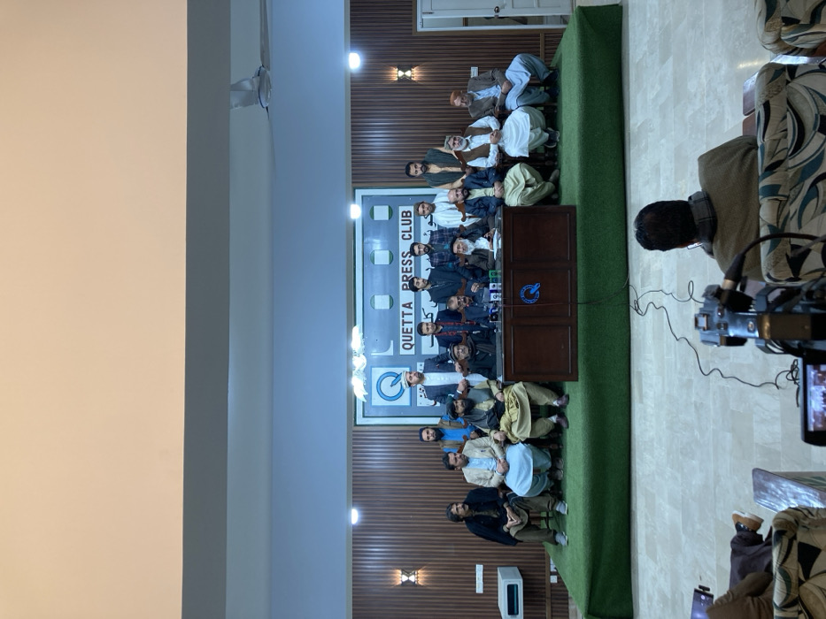 An indoor scene depicts a press conference or public address at the Quetta Press Club in Quetta City Tehsil, Pakistan. A group of eighteen adult males is arranged on a raised, green-carpeted platform against a wood-paneled wall. Five men are seated behind a dark wooden table with a "Q" logo, microphones, and small bottles, with one individual actively speaking into the microphones. Seven additional men are seated to the sides of this table in a semi-circular arrangement. Six younger men stand behind the seated individuals. All individuals on the platform are dressed in a mix of traditional Pakistani attire, including shalwar kameez, waistcoats, and various headwear. The wall behind them features a large white banner with a "Q" logo and the text "QUETTA PRESS CLUB" in English, along with Urdu script. Two decorative wall sconces are visible on the paneled wall. In the foreground, from behind, two individuals are seated in patterned chairs, one operating a video camera on a tripod, which is recording the event. Cables are visible on the tiled floor in the audience area. The ceiling features multiple recessed lights and a white ceiling fan. A white door is visible on the right side of the platform, and a white rectangular device is mounted on the left wall.