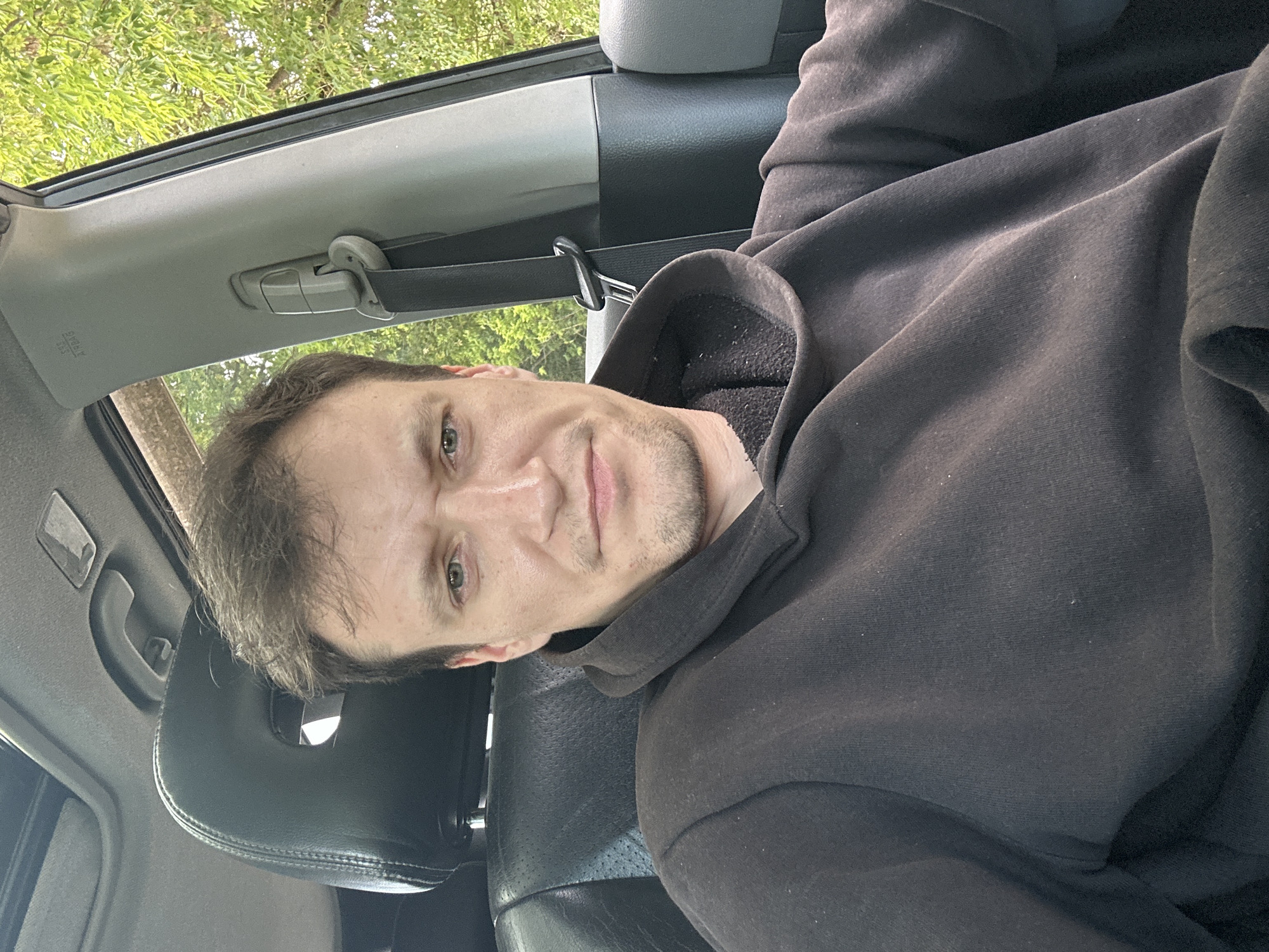 A male subject with light brown hair and a light beard is sitting in the driver's seat of a car. He is wearing a dark-colored hooded sweatshirt. The car has a black leather headrest and a sunroof. A seatbelt is visible on the right side of the frame. Outside the car, green trees are visible, suggesting an outdoor, possibly rural, setting. The image appears to have been taken during daylight. The location is identified as Makiv, Ukraine.
