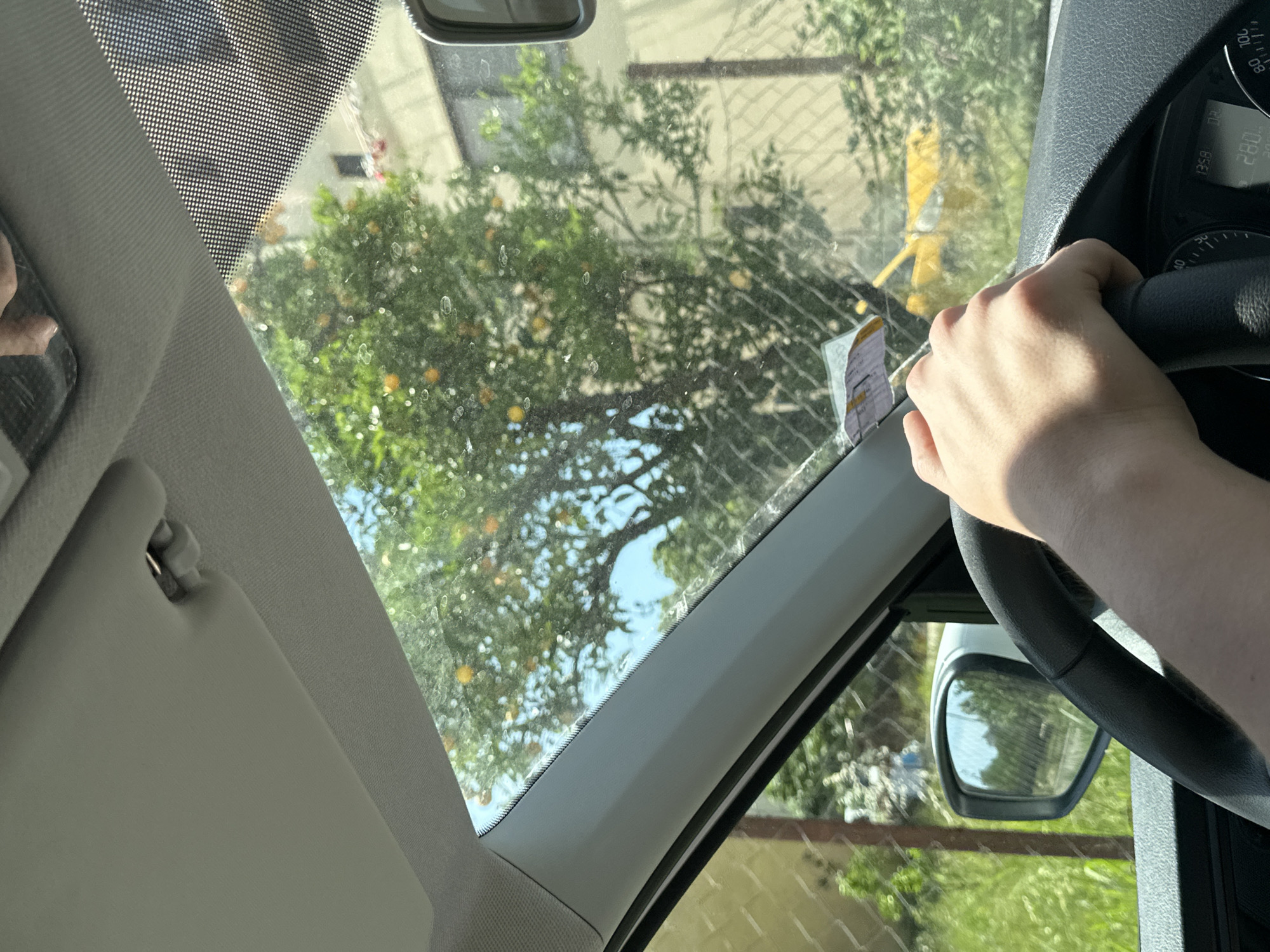 The scene is inside a vehicle, potentially in Bar, Montenegro, indicated by a vignette sticker visible on the windshield. The vehicle is stationary or moving at a slow speed, as the hand is gripping the steering wheel. Outside, a tree with round yellow fruit is visible through the windshield, along with a fence and buildings, suggesting an urban or suburban environment. The sun visor is down on the left side. The car's speedometer is visible, indicating a speed of 0 km/h. The time on the dashboard indicates it is 13:58.