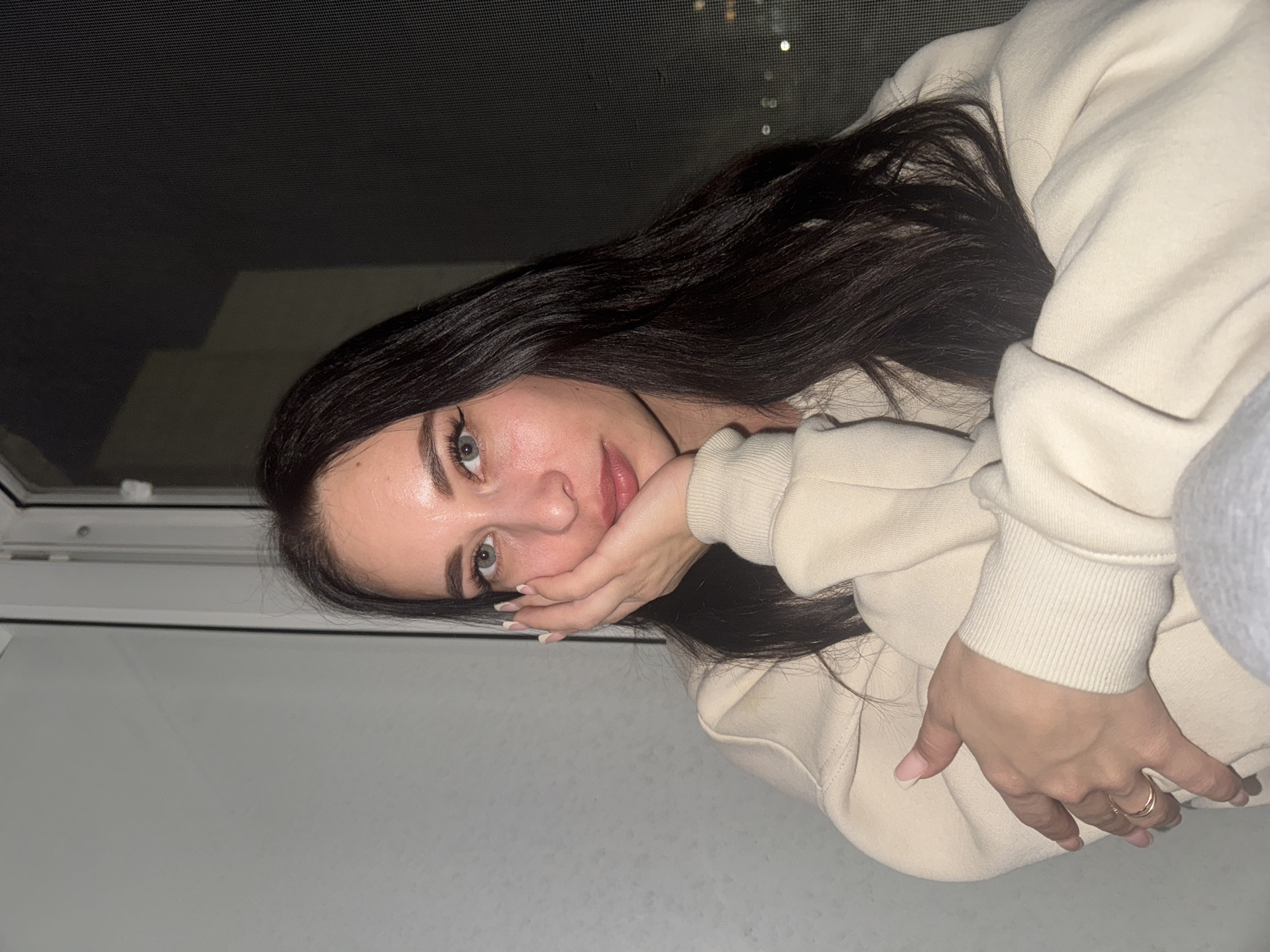 A young woman with long, dark hair and light eyes is posing for a photograph. She is wearing a light-colored sweatshirt and has her hand near her face. A window with a dark mesh screen is visible in the background, and lights from the city of Chaikovsky, Russia, can be seen through the screen.