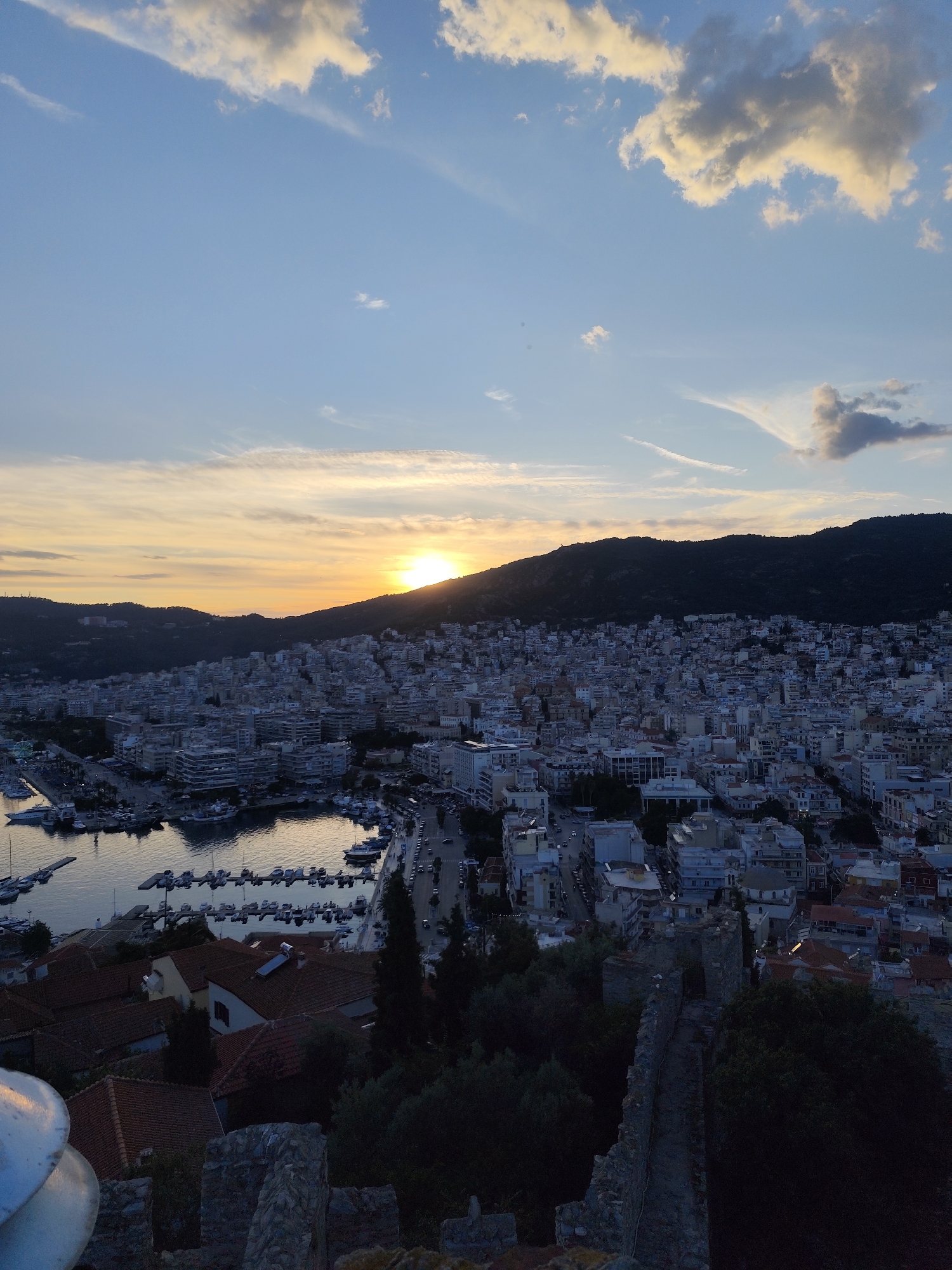 An aerial, full shot captures a city, likely Kavala, Greece, at sunset. The sun dips behind a dark, tree-covered mountain range that forms a silhouette against the bright, orange-yellow horizon. Below the mountain, a dense urban landscape of multi-story buildings with white and grey facades stretches towards a harbor filled with numerous docked boats. The sky above is a gradient of blues and whites, with scattered clouds. In the foreground, the stone ruins of a castle wall are visible, casting a shadow over lush green trees. The overall scene is one of a coastal city at dusk.