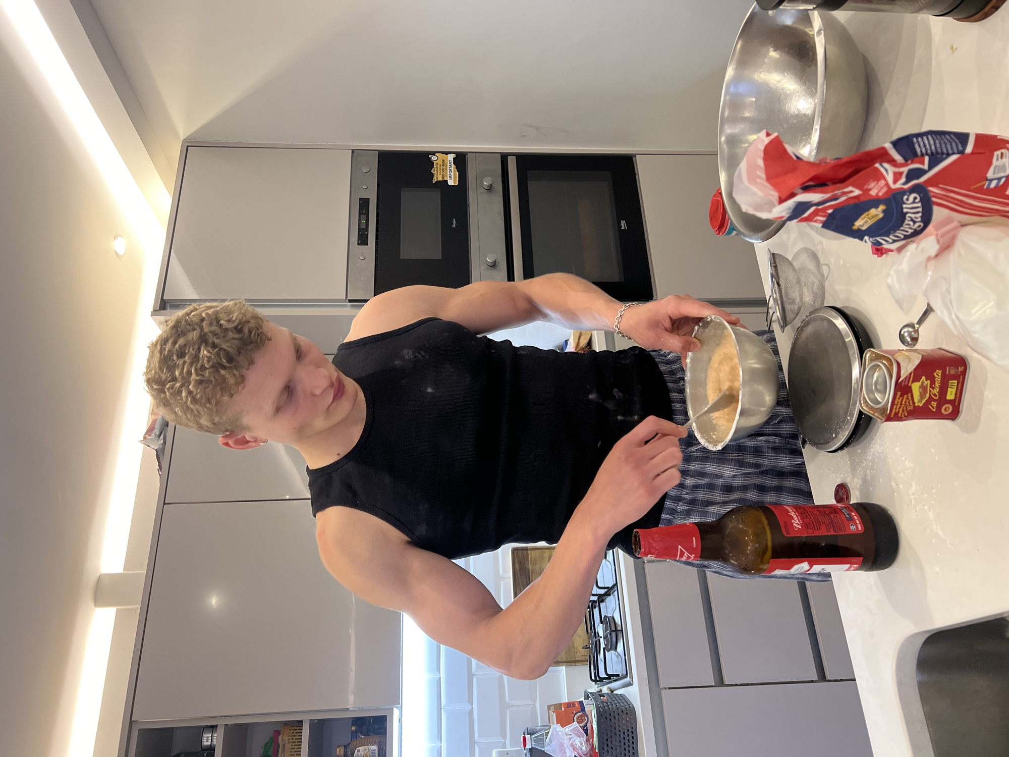 A young Caucasian man with curly blonde hair, wearing a black tank top and checkered pajama bottoms, stands in a modern kitchen and appears to be mixing ingredients in a metal bowl. He is in the process of cooking or baking. The kitchen has light grey cabinets and countertops, as well as stainless steel appliances including a microwave and oven. On the counter, there is a bottle of Budweiser beer, a can of "La Chinata" paprika, a small metal sifter, a digital scale, and a large mixing bowl. A bag with a Union Jack pattern is also visible. The overall setting suggests a domestic environment in London, United Kingdom.