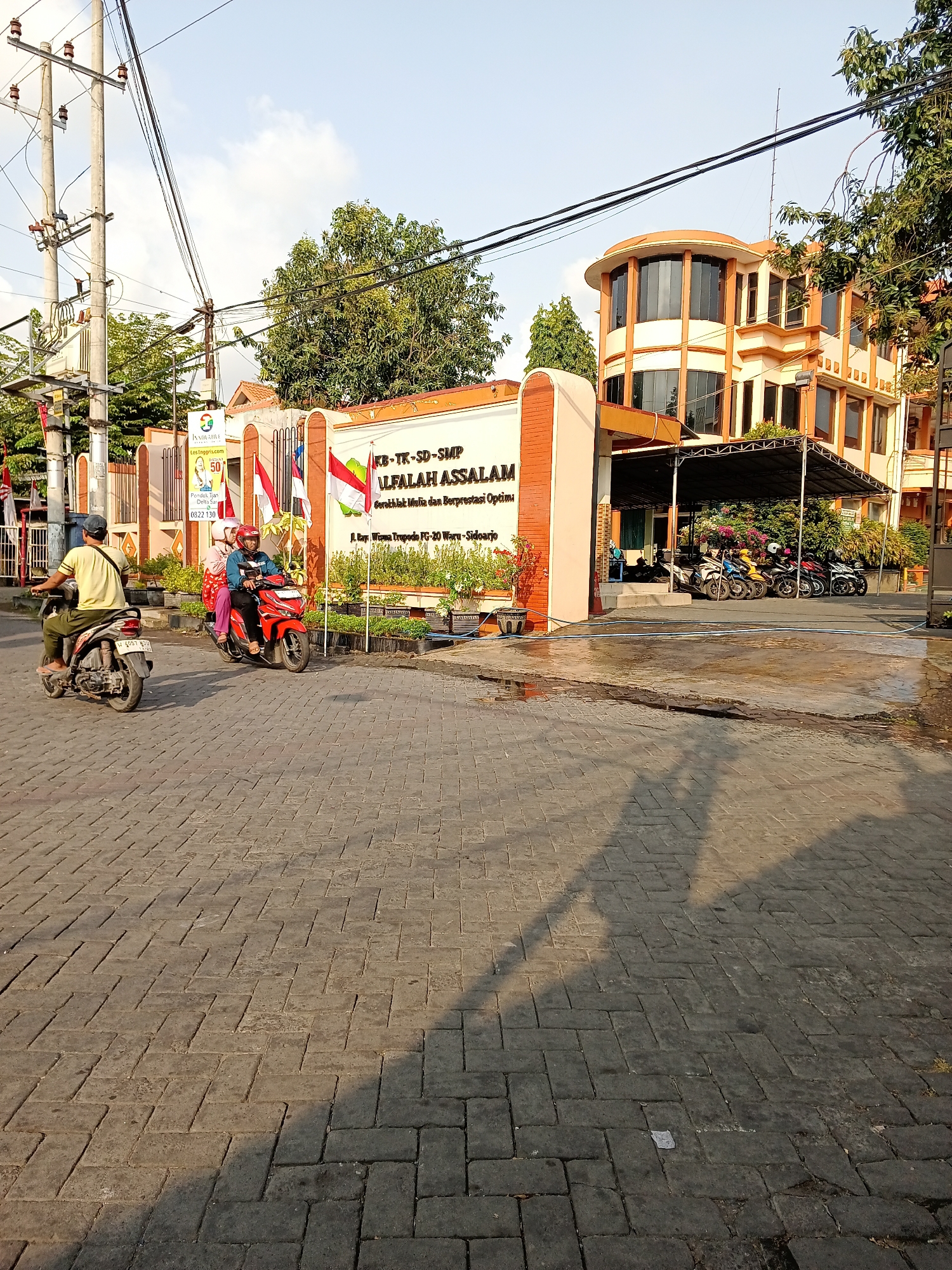 This is a medium shot, captured during daylight hours, of the street in front of an educational institution in Tropodo, Indonesia. The foreground is dominated by a patterned brick road, reflecting the warm, late afternoon sun. In the middle ground, two motorbikes are visible. The first, a silver scooter, is ridden by a person wearing a yellow t-shirt and a blue cap, moving away from the camera. Behind it, a red scooter carries two individuals, both wearing helmets, traveling in the same direction.
The background features the facade of the educational institution, identified by the sign "KB-TK-SD-SMP ALFAALAH ASSALAM" and an address below it, which, based on the visual context and common knowledge of the area, is likely located in Tropodo, Sidoarjo Regency, East Java, Indonesia. The building is a multi-story structure with orange and white accents, featuring prominent rounded windows on the upper floor. A covered parking area with several motorbikes is visible to the right of the entrance. The entrance itself is flanked by a fence and some decorative potted plants, and Indonesian flags are displayed prominently. Overhead, power lines crisscross the sky, and mature trees provide shade on the right side of the frame. The overall atmosphere is that of a typical urban street scene in Indonesia, with a clear emphasis on the educational facility.