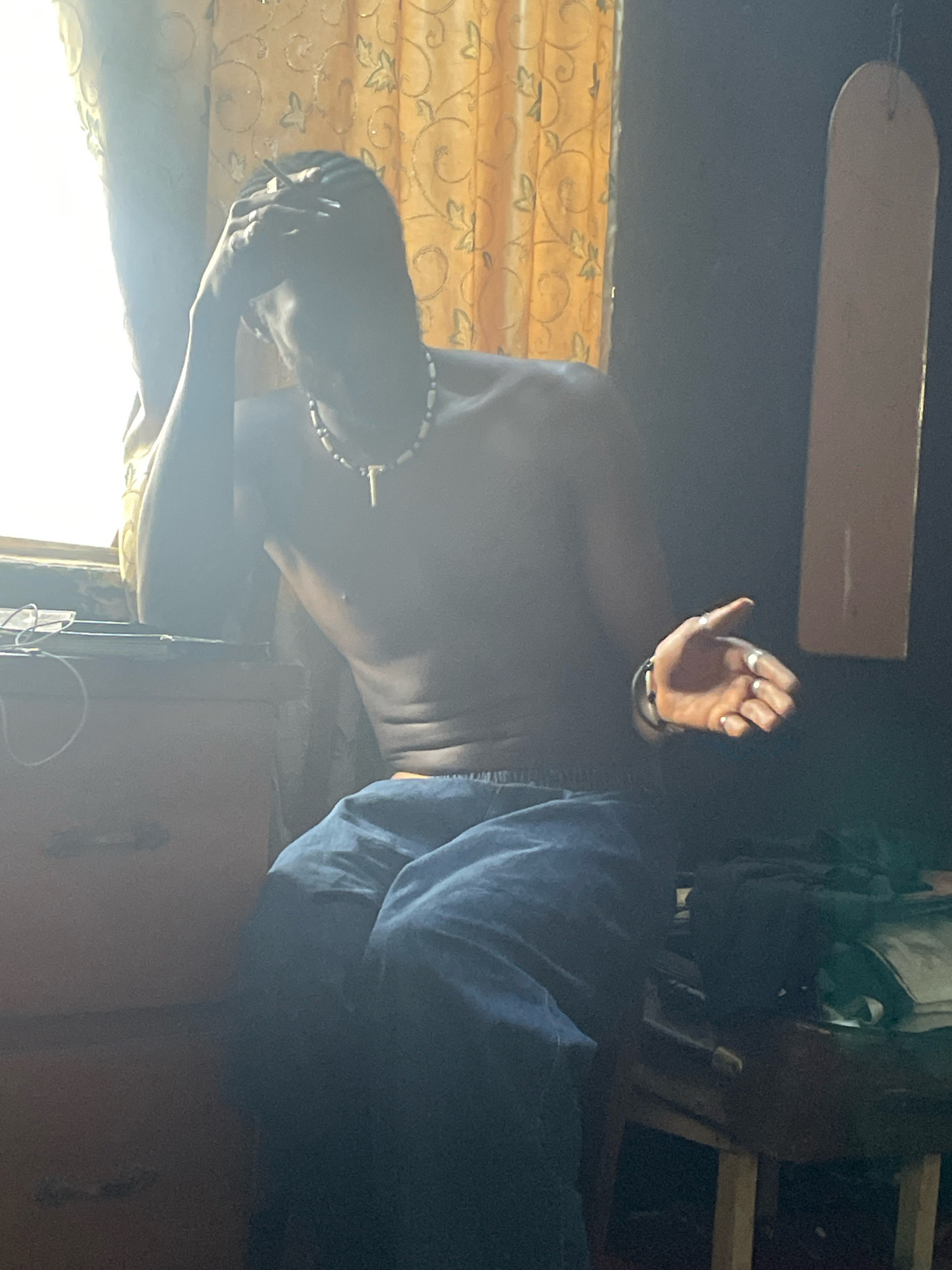 A shirtless Black male is seated indoors, leaning against a window frame, with his head in his hand, appearing distressed. He is wearing blue jeans and a beaded necklace with a cross pendant. To his left, a dresser is visible, and to his right, a wooden table holds various items, including a long, light-colored object leaning against the wall. The room is dimly lit with yellow patterned curtains visible behind the subject. The scene appears to be an interior residential space, potentially in Jos, Nigeria, given the context provided, though no specific indicators of location are visually apparent within the image itself.